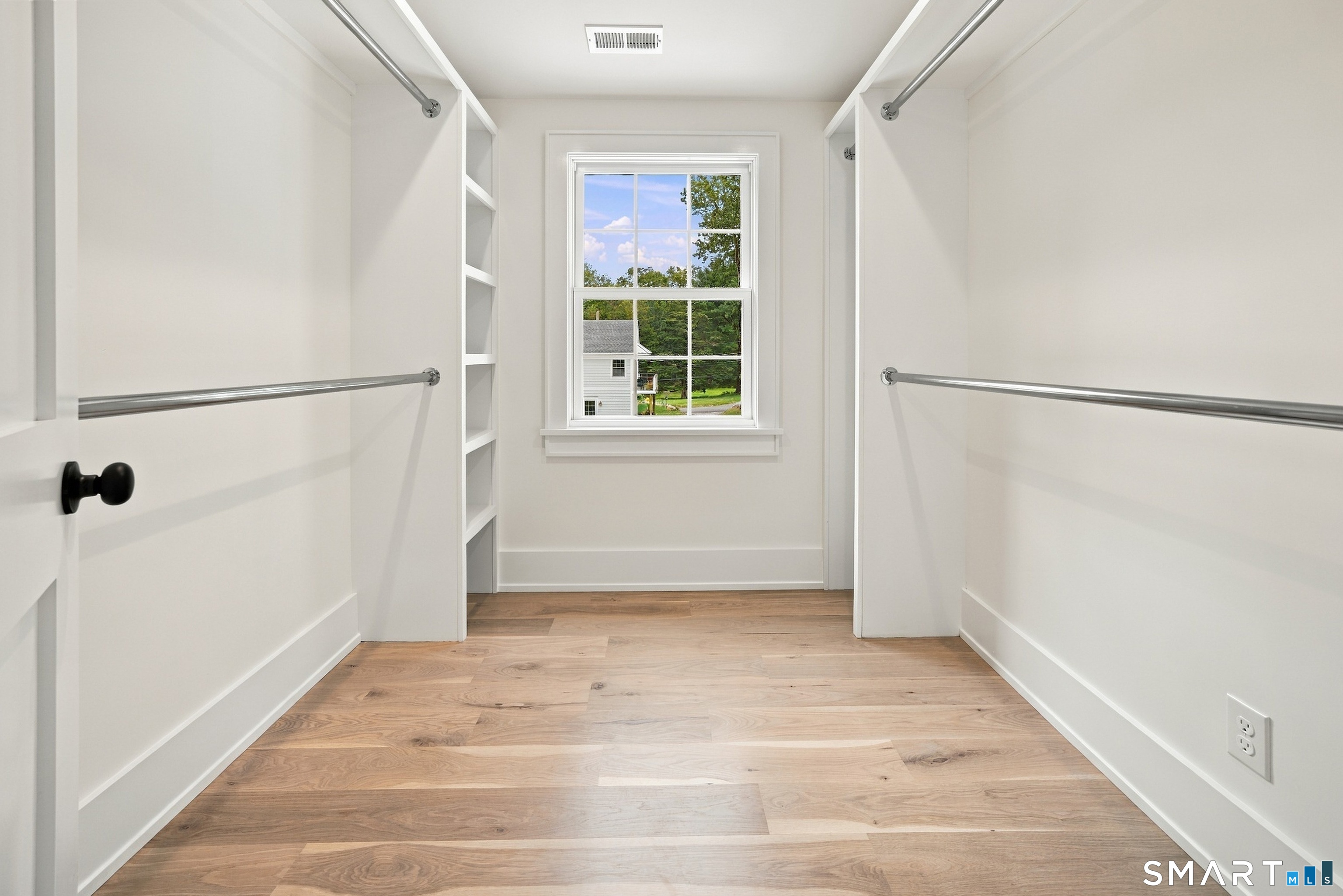 Walk-in closet with built-in shelving, two hanging rods, hardwood floors, and a window overlooking trees.