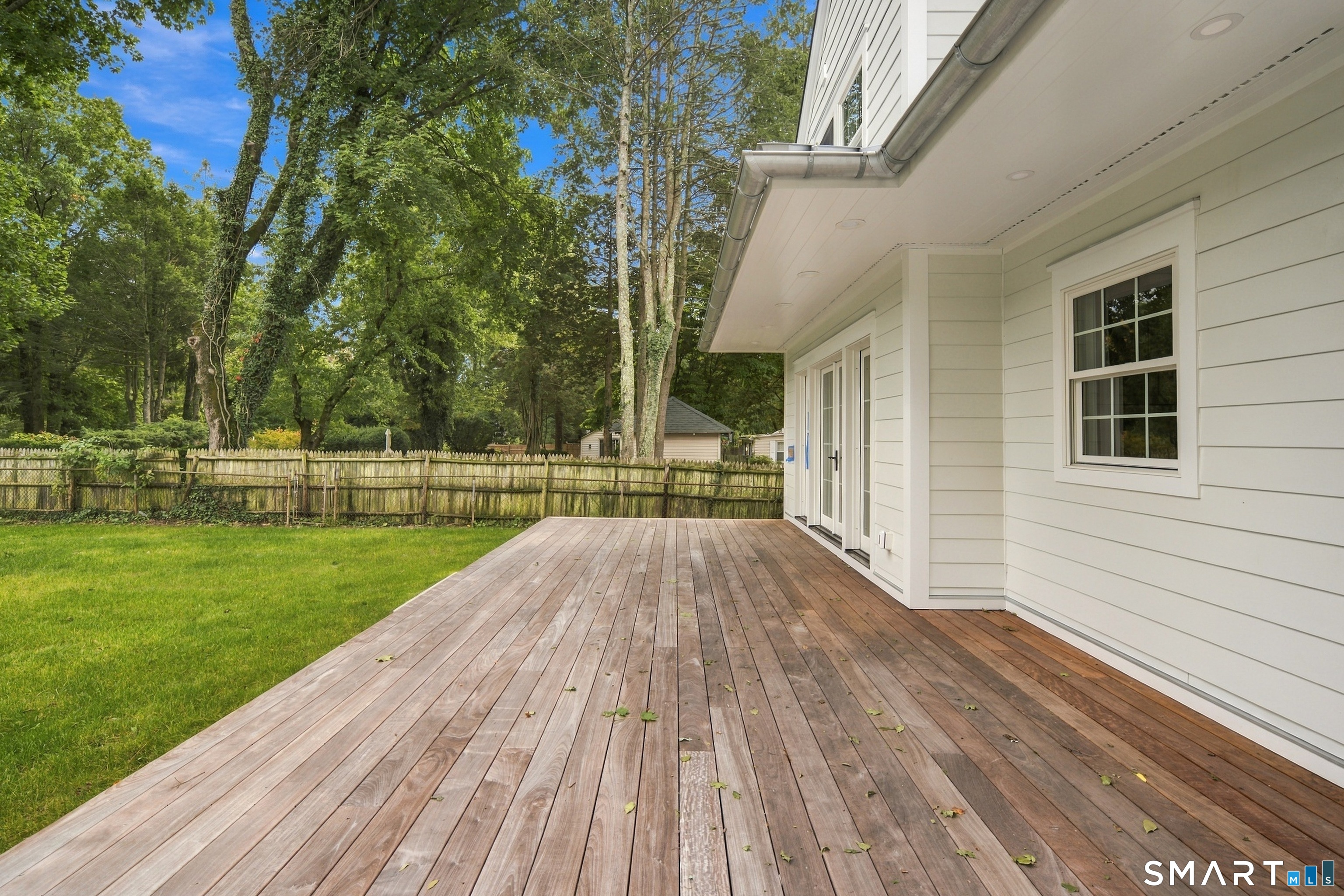 Wooden deck beside the white house overlooks a fenced lawn and mature trees.