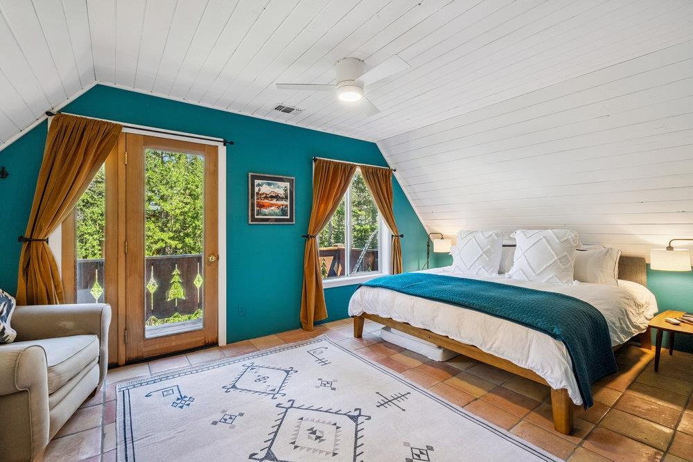 Bedroom with a teal accent wall, sloped ceiling, and wood-framed glass doors opening to trees.
