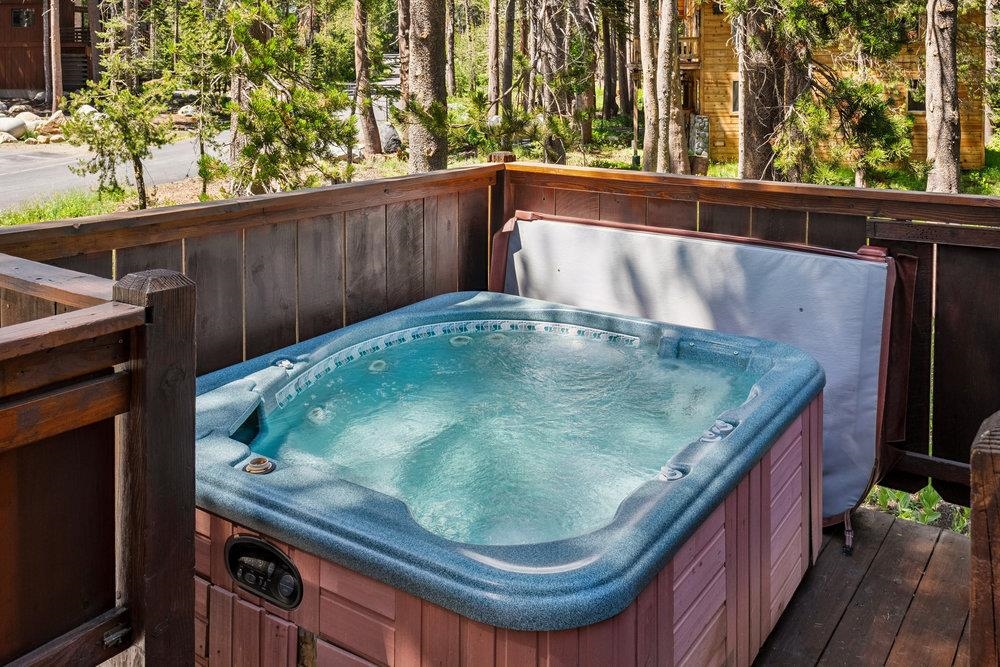 Outdoor hot tub on a wooden deck, surrounded by evergreen trees and cabins.