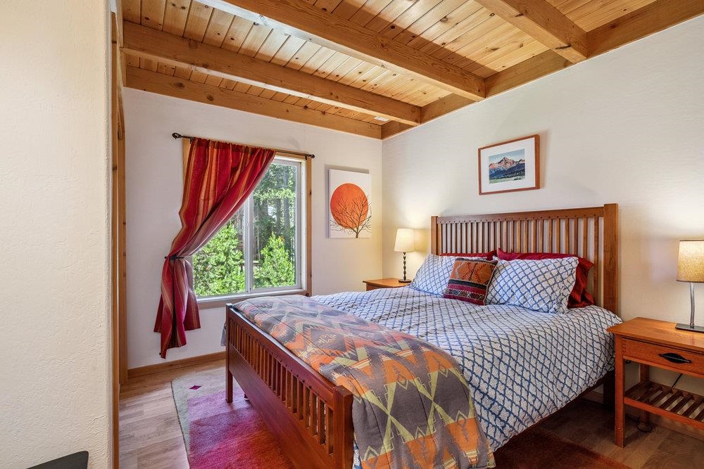Bedroom with a wooden bed beneath a vaulted plank ceiling and a window overlooking trees.