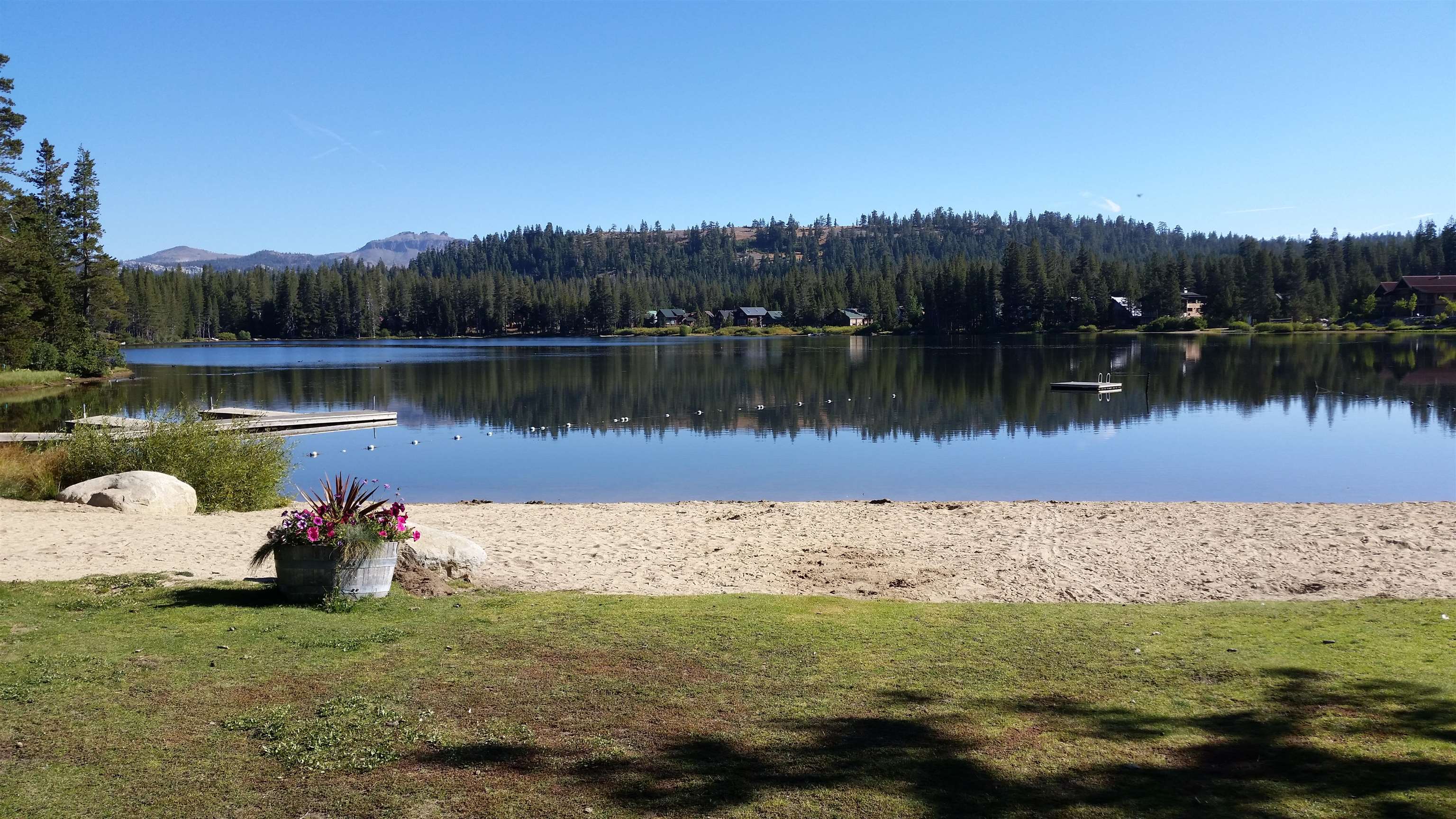 Sandy lakeside beach and dock beside a calm lake backed by pine-covered hills.