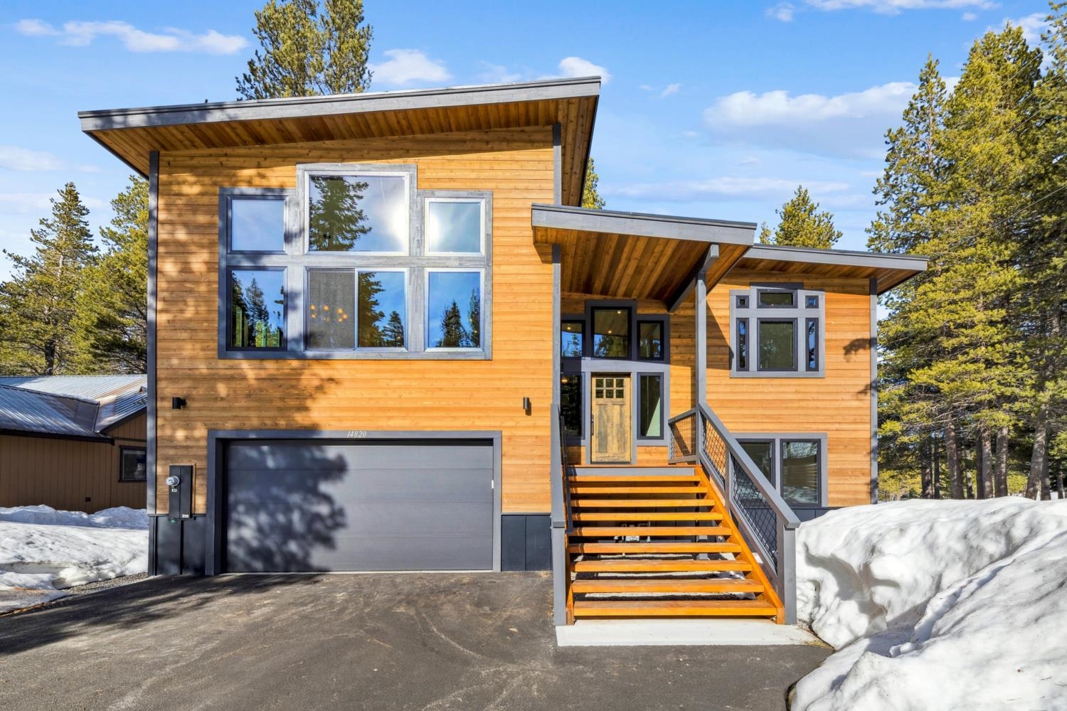 Modern wood-sided home with a gray garage and orange-toned entrance stairs, surrounded by snow.