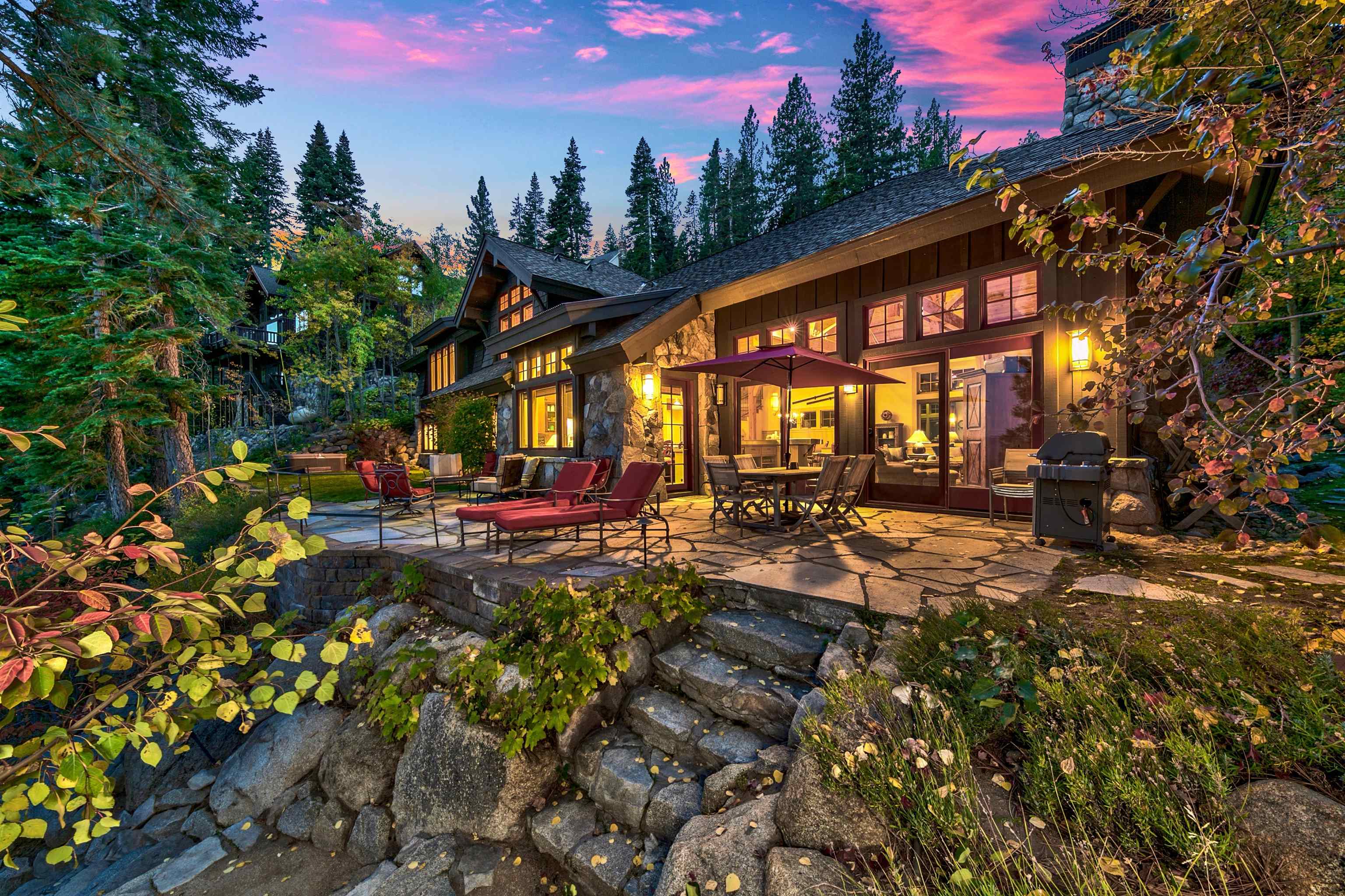 Rustic mountain home with a stone patio, outdoor dining set, and glowing windows at dusk.