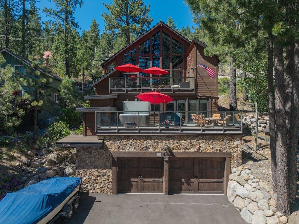 Pine-wooded home with glass-railed decks, red umbrellas, stone retaining wall, and two-car garage.