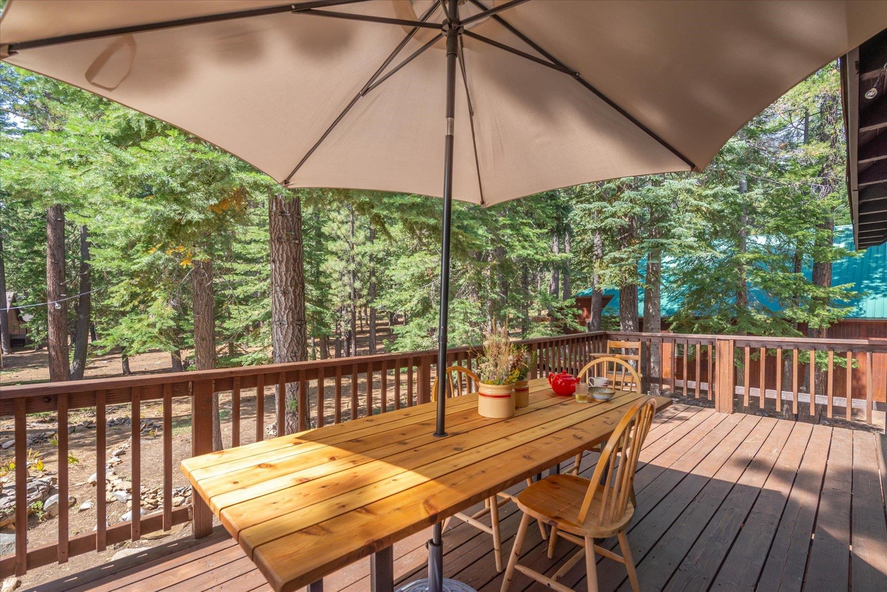 Wooden deck dining table beneath a large umbrella, overlooking a wooded yard.