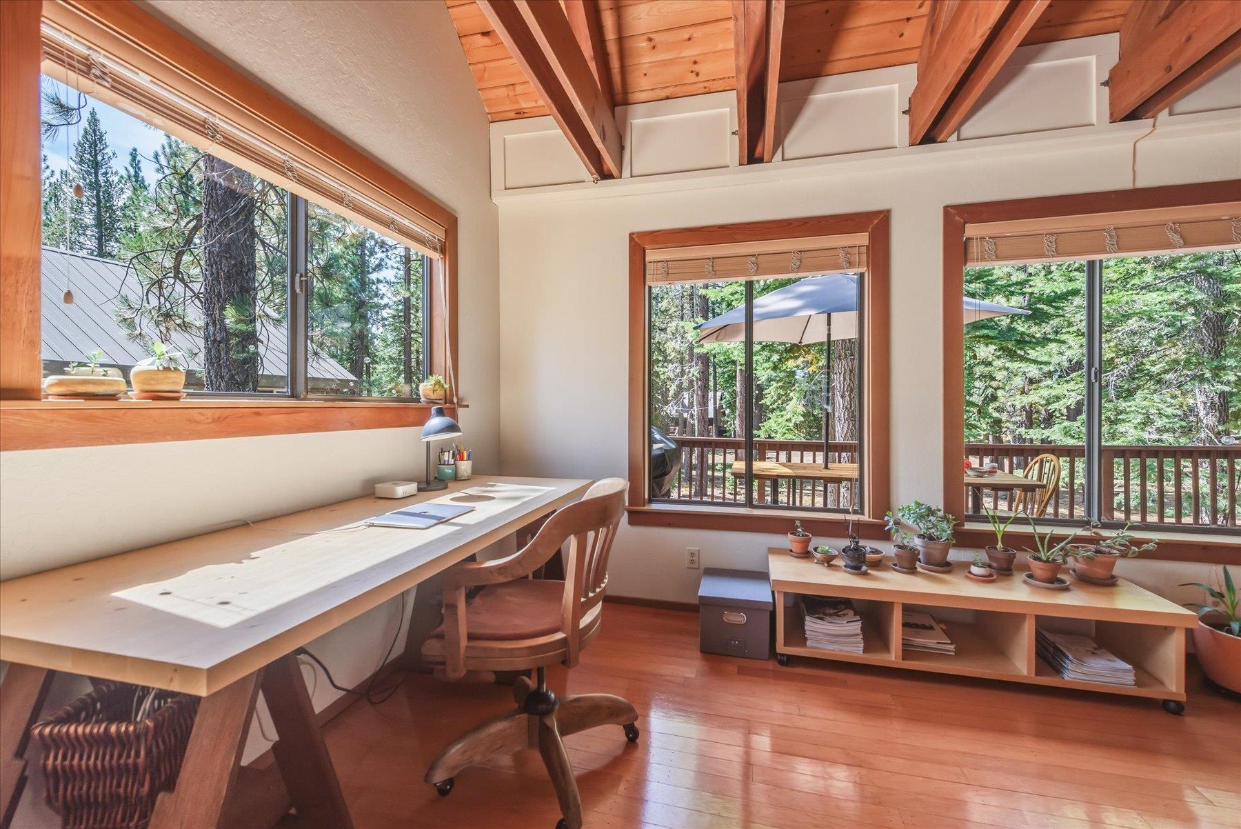 Home office with exposed timber beams, a long desk, and windows overlooking trees and a deck.