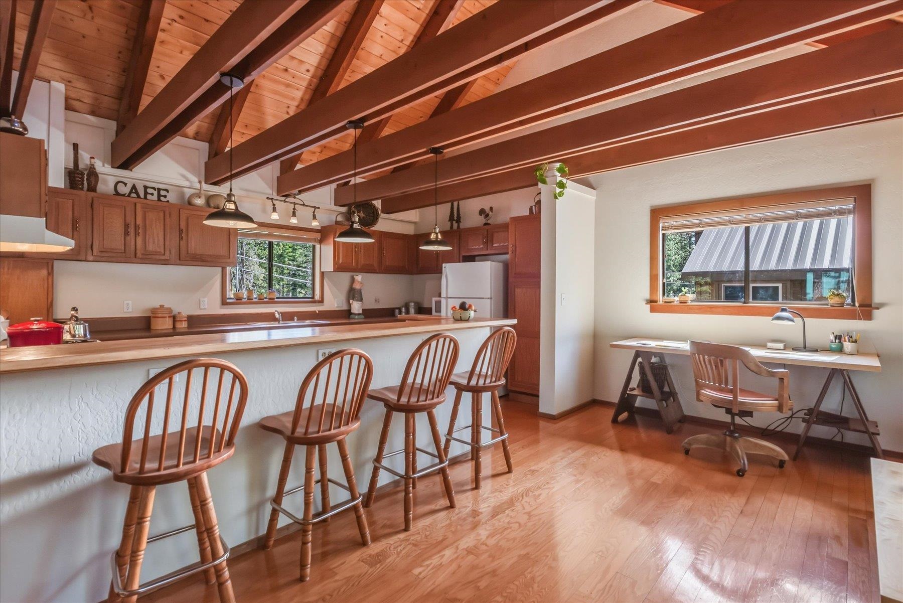 Kitchen with a long breakfast bar, wooden stools, exposed ceiling beams, and an adjacent desk by the window.