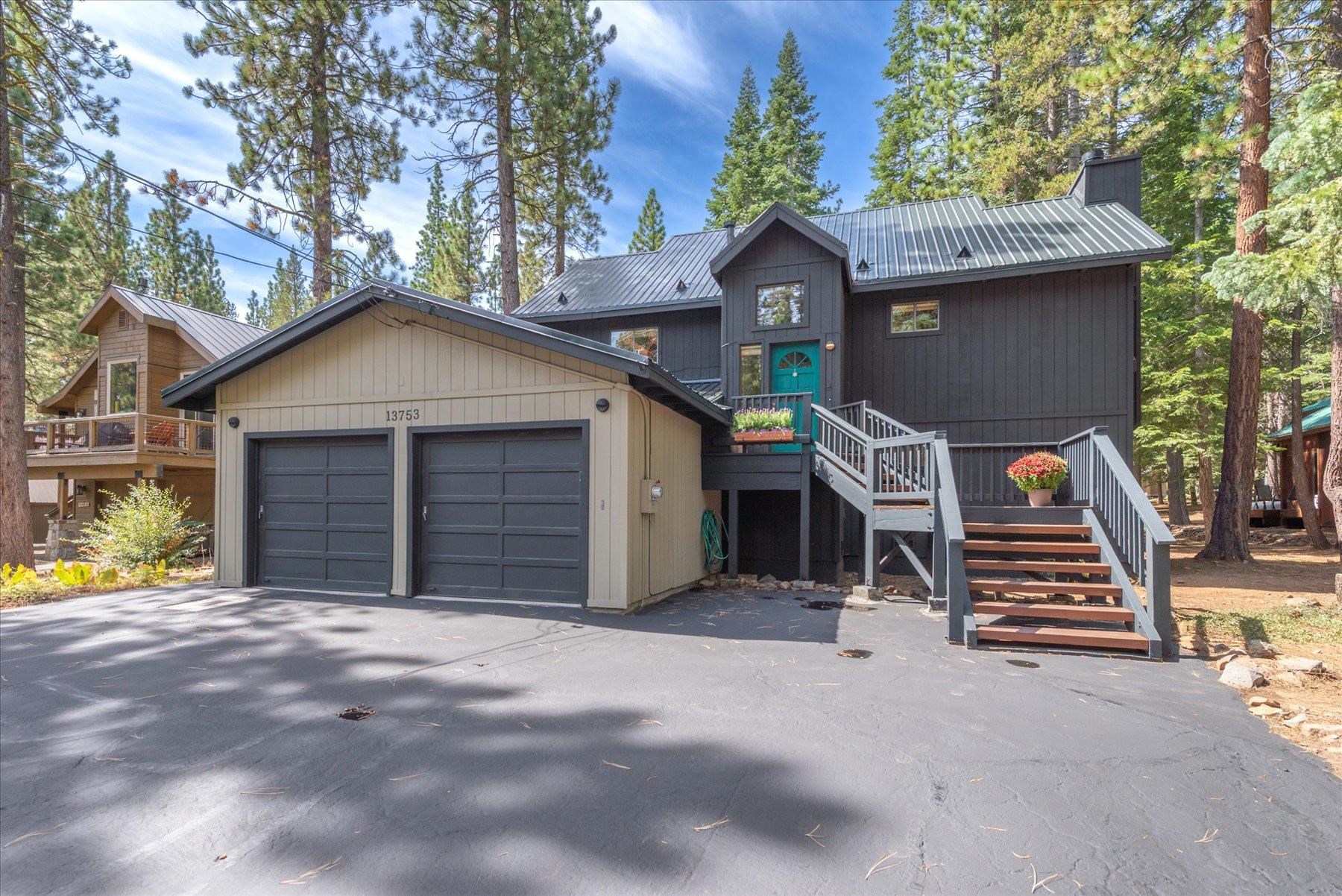 Dark-sided home with a turquoise door, raised wooden stairs, and attached two-car garage among pine trees.