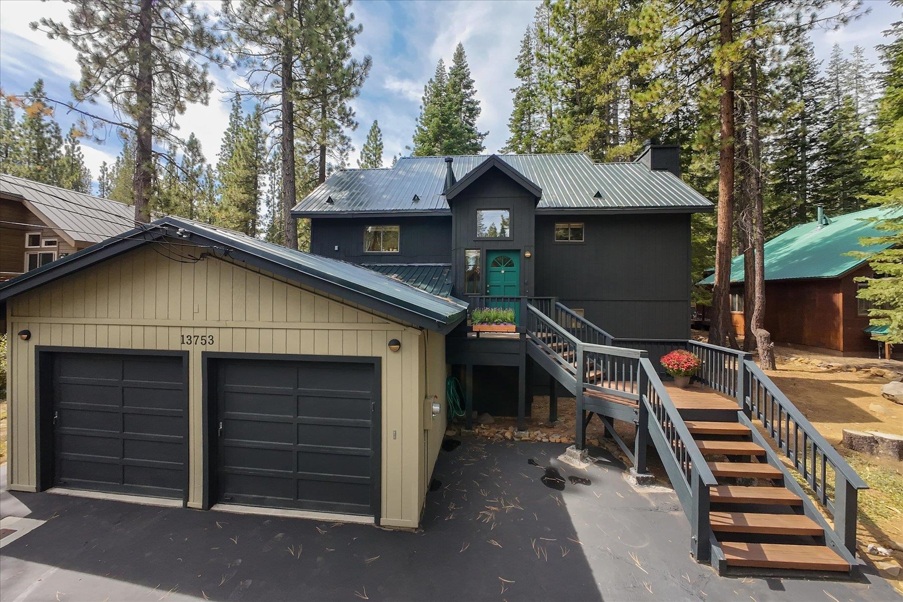 Dark charcoal house with teal door and raised entry stairs beside a two-car garage among pine trees.