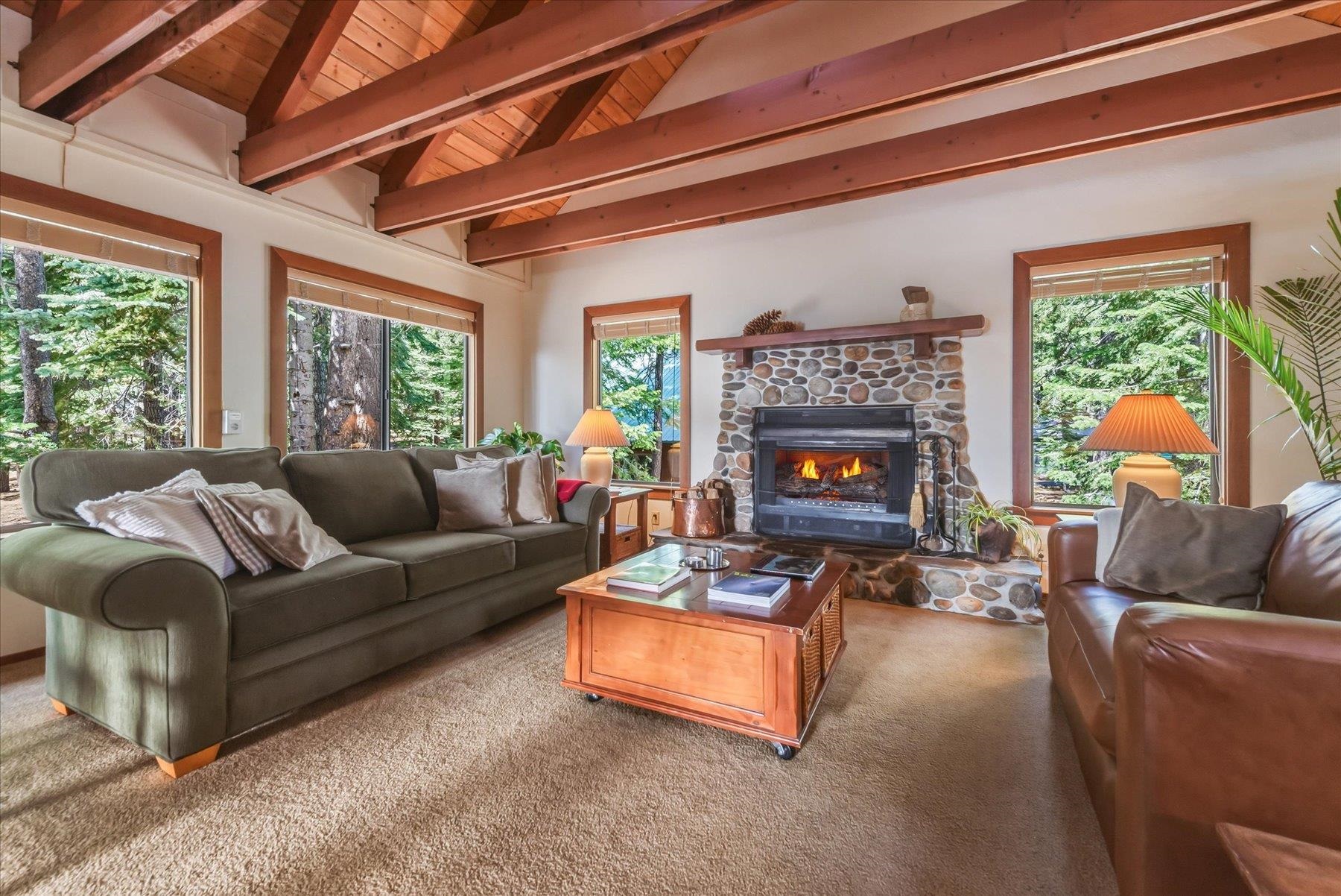 Living room with exposed wood beams, a stone fireplace, and large windows overlooking trees.