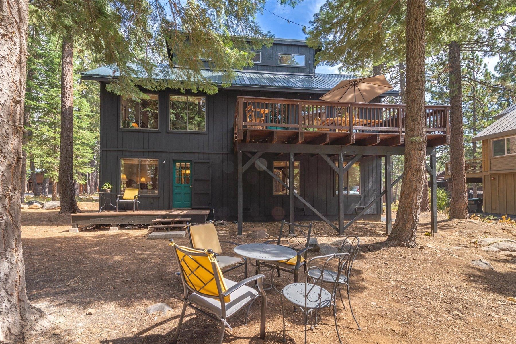 Dark two-story home with a raised deck and patio seating among tall pines.