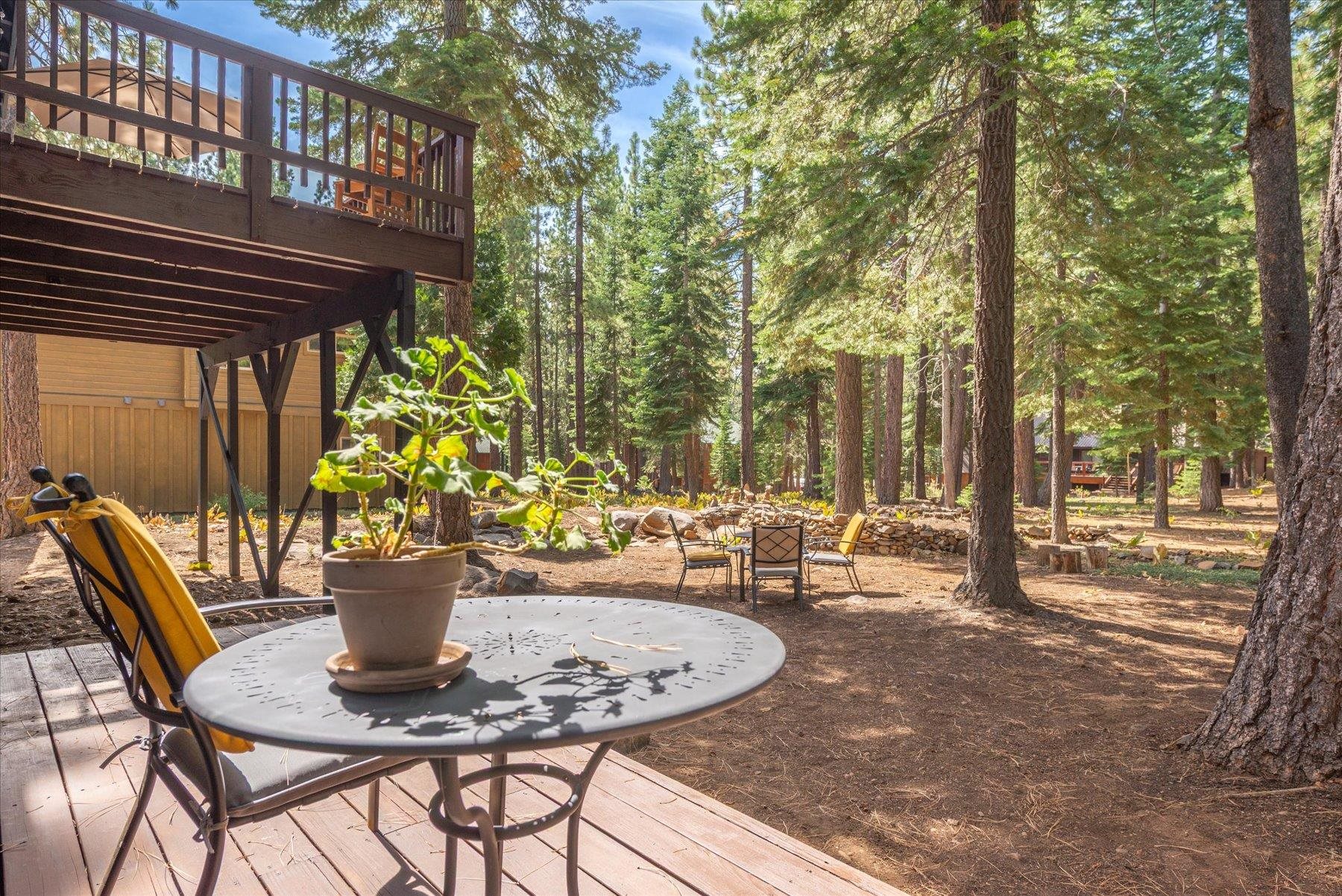 Outdoor patio with a round table beneath an elevated deck, surrounded by tall pine trees.