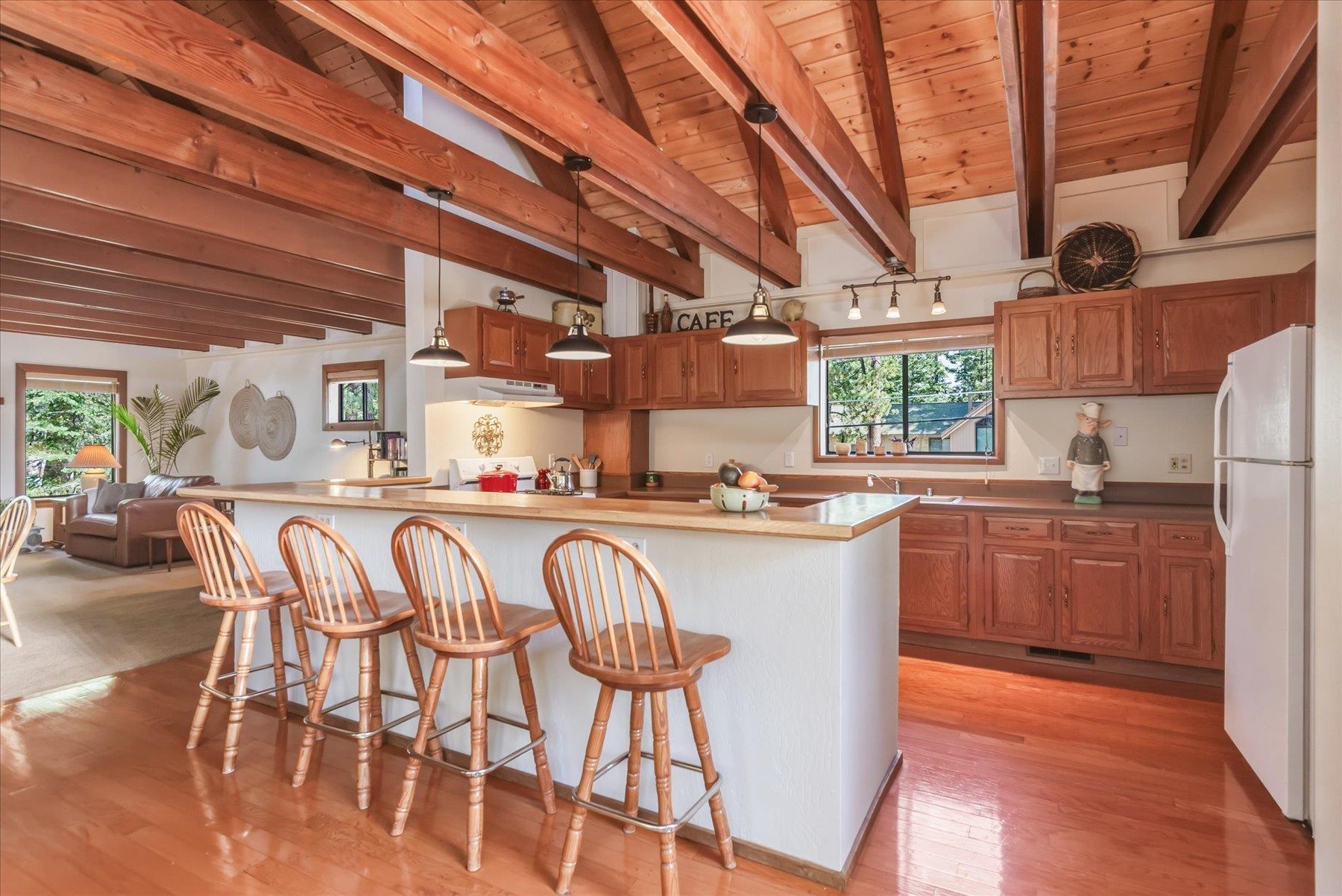 Open kitchen with a white island, wooden bar stools, reddish cabinets, and exposed timber ceiling beams.