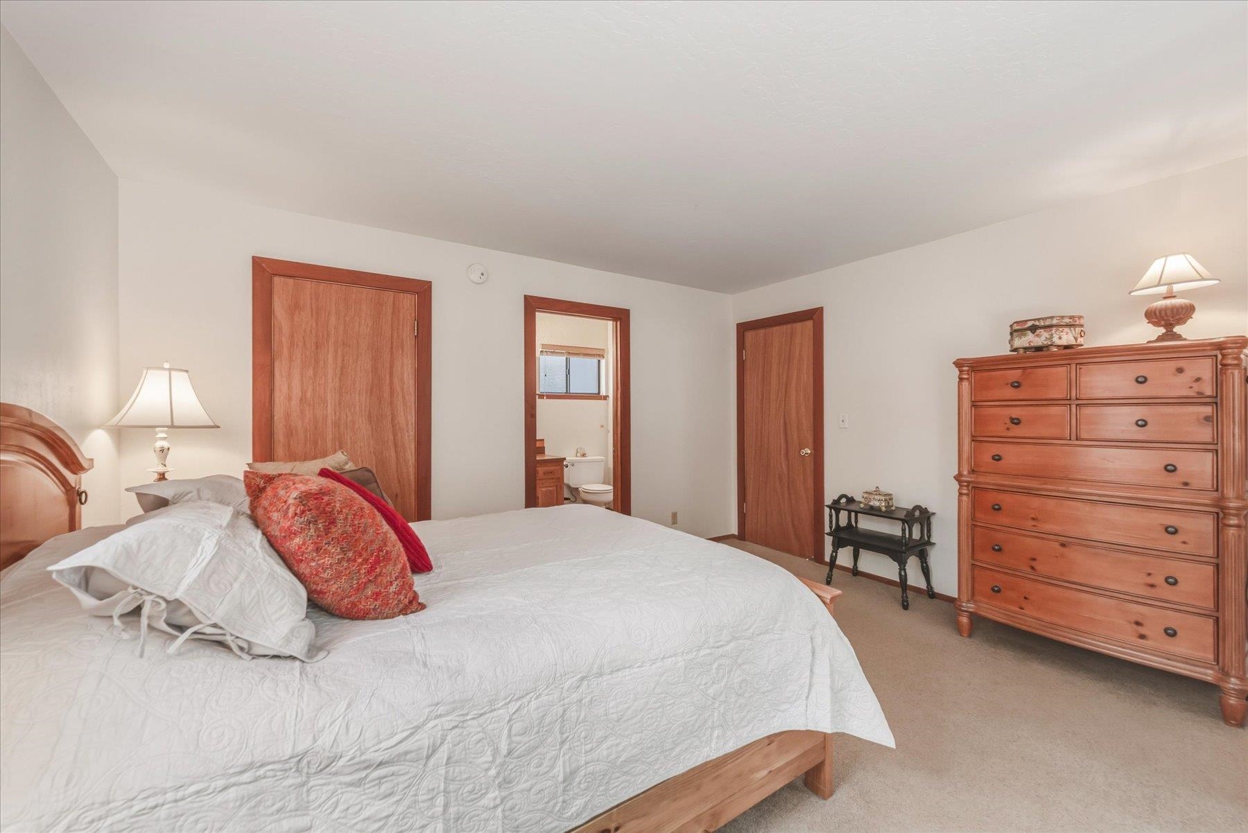 Furnished bedroom with a white-quilted bed, reddish wood dresser, and bathroom visible through the doorway.