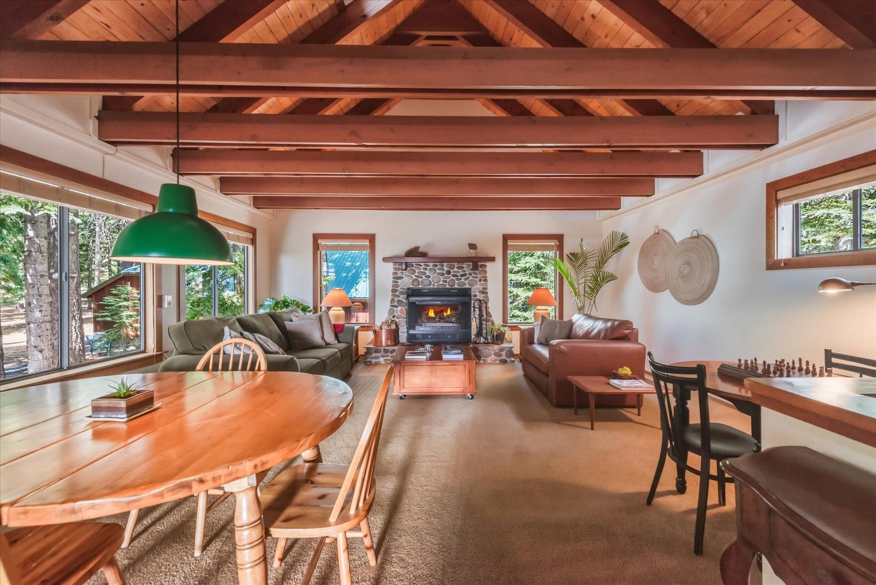 Living room with exposed wood beams, a stone fireplace, and large windows overlooking trees.