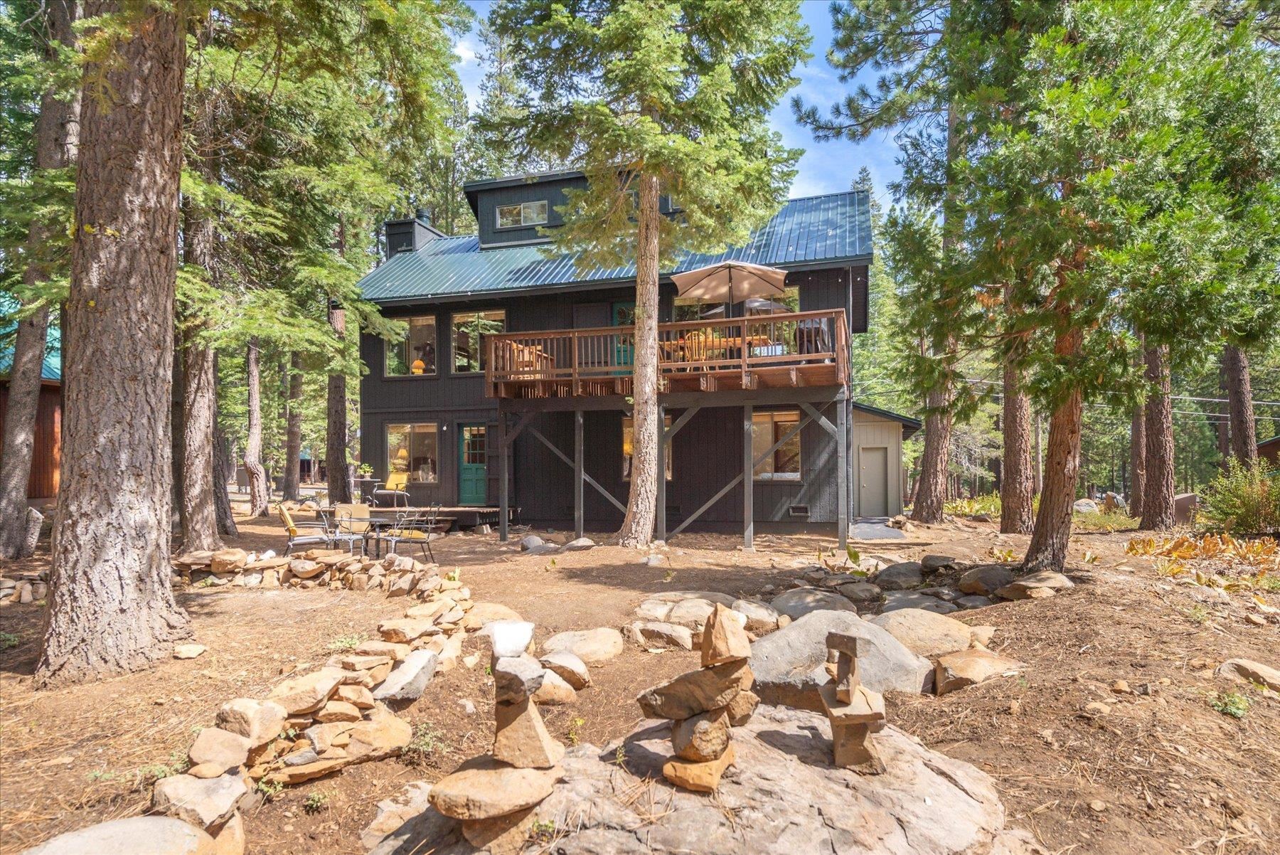 Dark two-story house with a raised wooden deck among pine trees and stone landscaping.