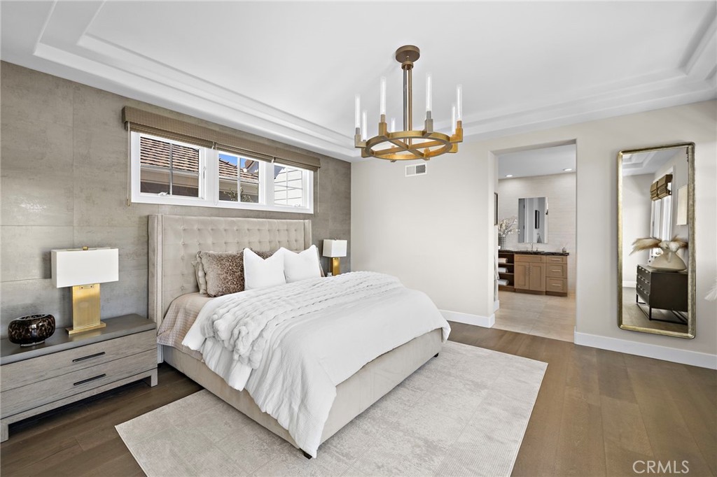 Bedroom with a white bed, gray upholstered headboard, brass chandelier, and adjoining bathroom vanity.