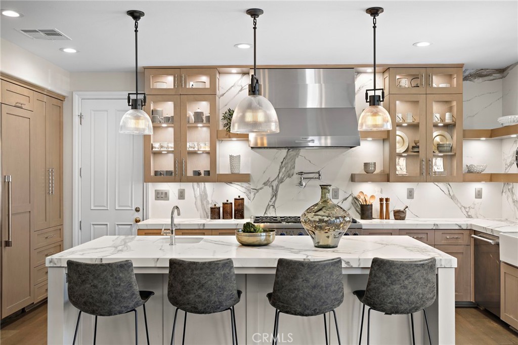 Kitchen with a large marble island, four gray stools, light wood cabinets, and a stainless steel range hood.