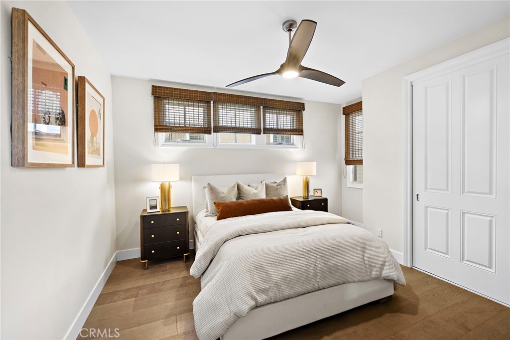 Bedroom with white bedding, woven window blinds, dark bedside tables, and wood flooring.