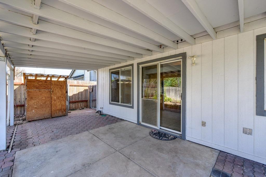 Covered patio with white roof beams, paver flooring, sliding glass door, and wooden shed.