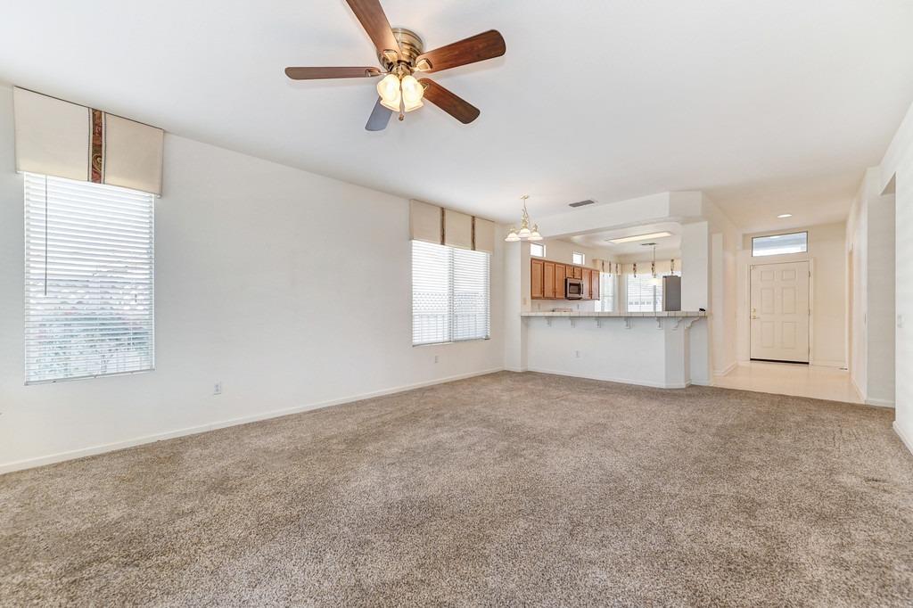 Empty carpeted living room with a ceiling fan and an open kitchen with breakfast bar.