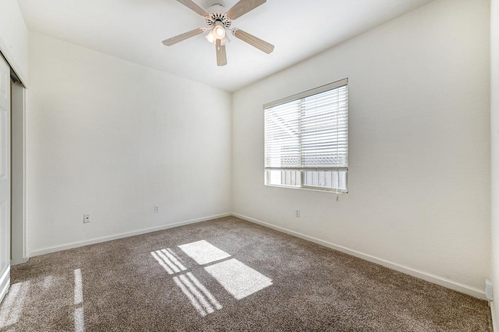 Empty bedroom with beige carpet, ceiling fan, and sunlight streaming through window blinds.