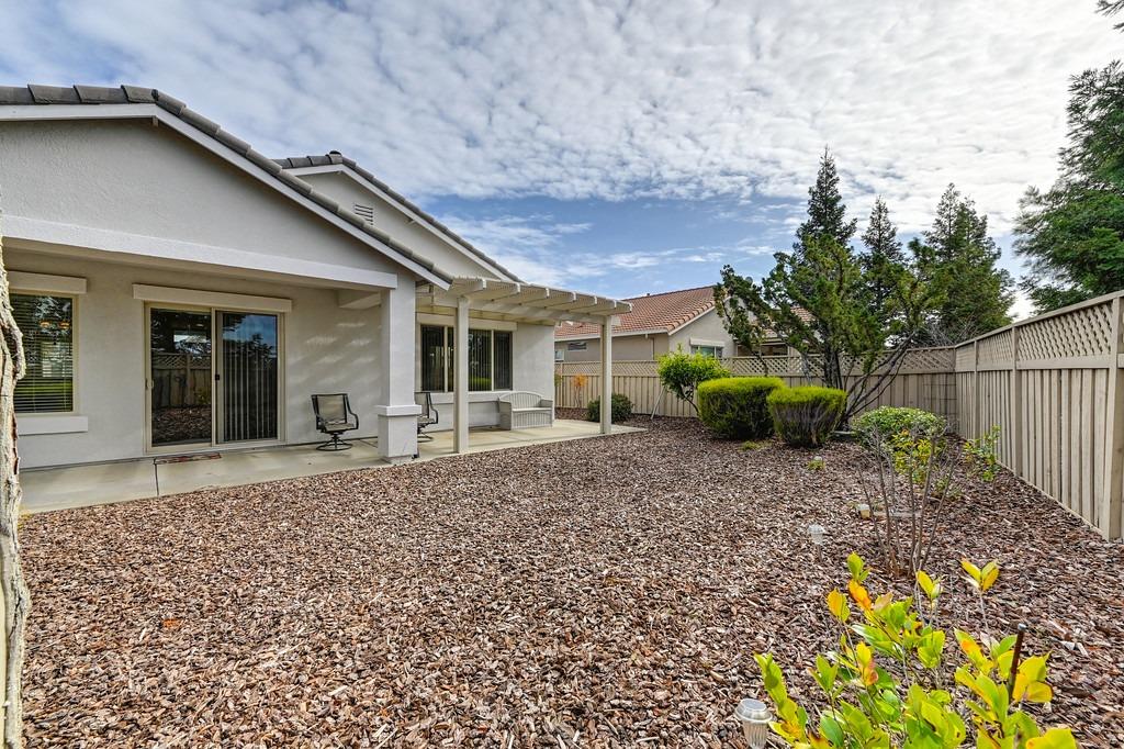 Home’s covered rear patio opens onto a fenced, bark-mulched yard with shrubs and small trees.
