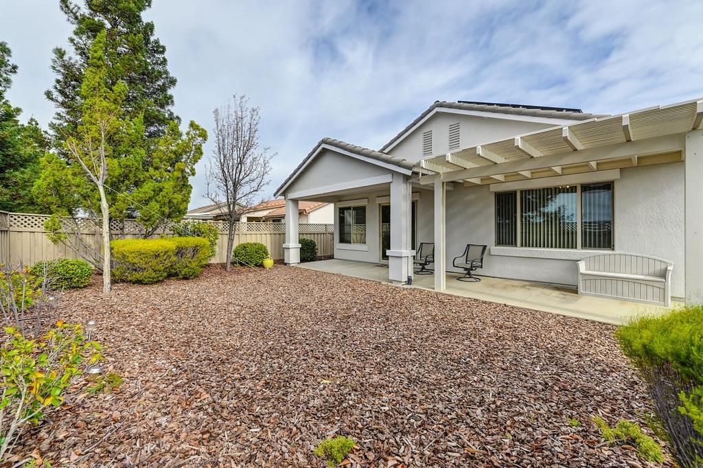 Home’s rear patio with a white pergola, outdoor chairs, and a wood-chip landscaped yard.