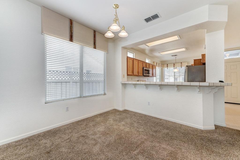Empty dining area with beige carpet, a three-light chandelier, and a pass-through counter to the kitchen.