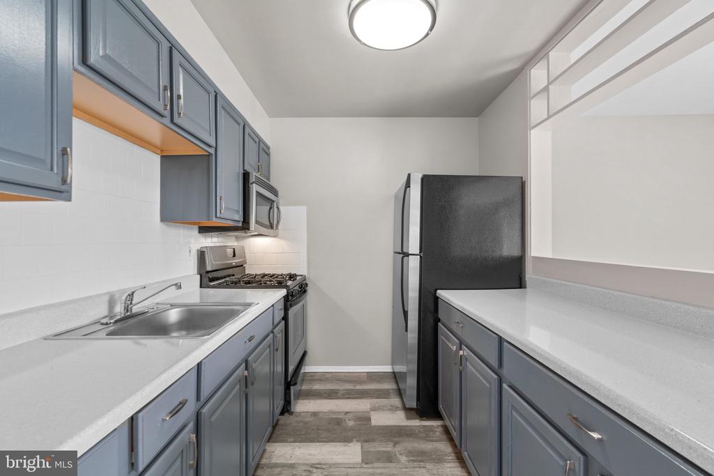 Galley kitchen with blue-gray cabinets, white counters, stainless-steel appliances, and wood-look flooring.