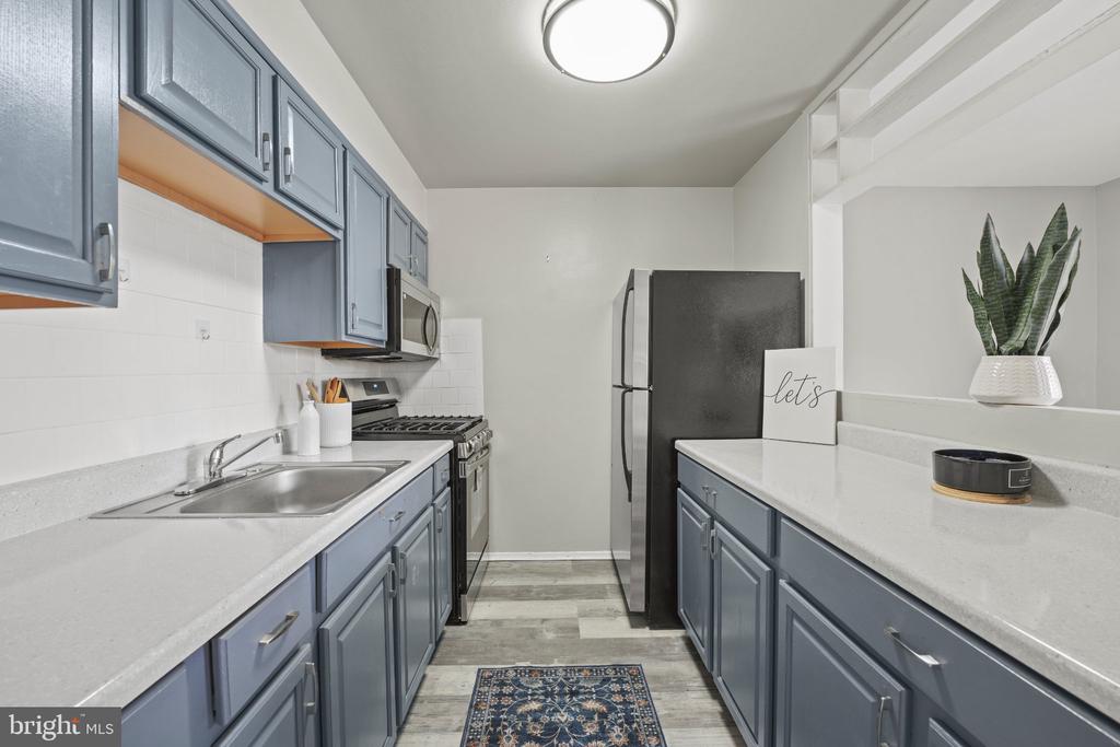 Galley kitchen with blue-gray cabinets, white counters, stainless steel appliances, and a patterned rug.
