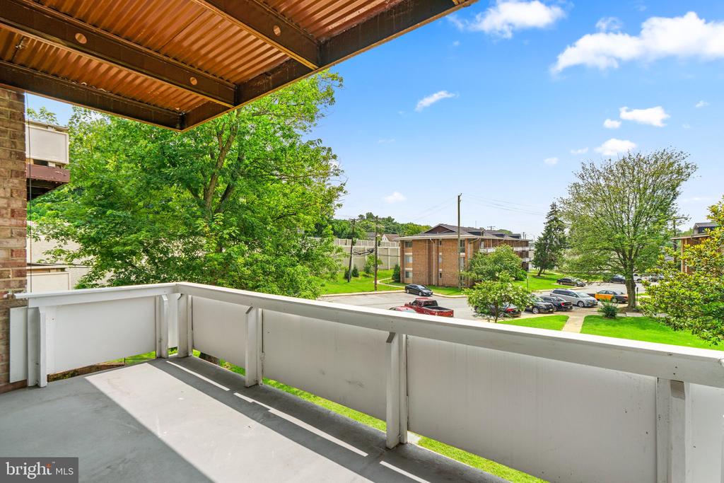 Covered apartment balcony overlooking leafy grounds, parked cars, and neighboring brick buildings.