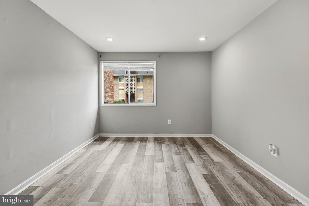 Empty room with gray wood-look flooring, recessed lights, and a window facing a neighboring building.