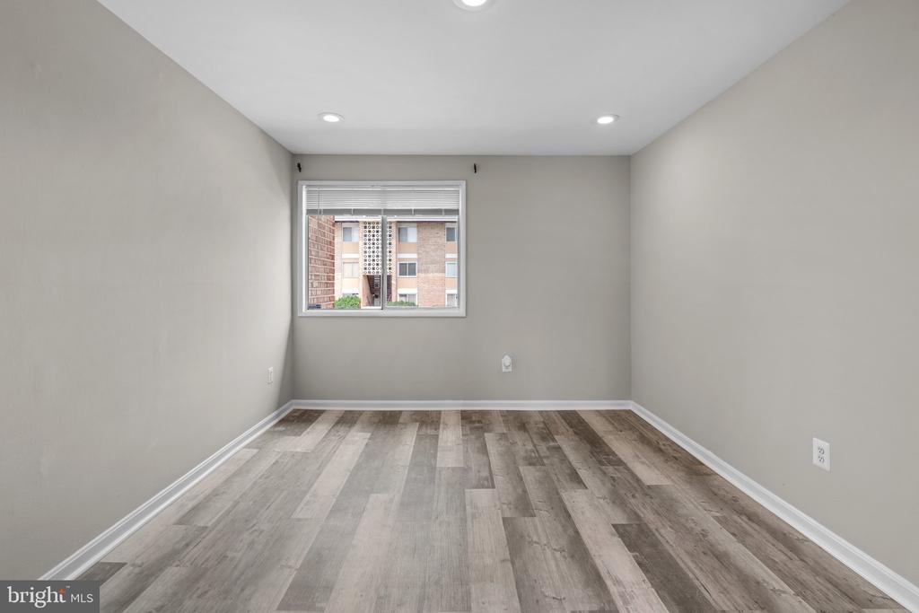 Empty room with gray walls, plank flooring, recessed lights, and a window overlooking a neighboring building.