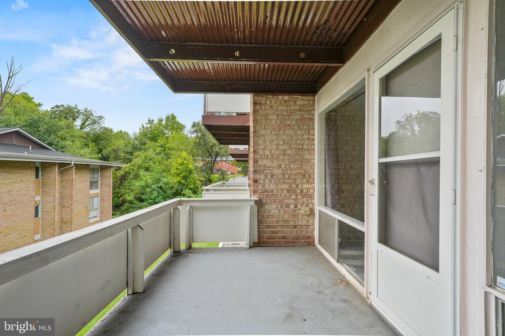 Covered balcony with a concrete floor, white railing, and views of trees and neighboring buildings.