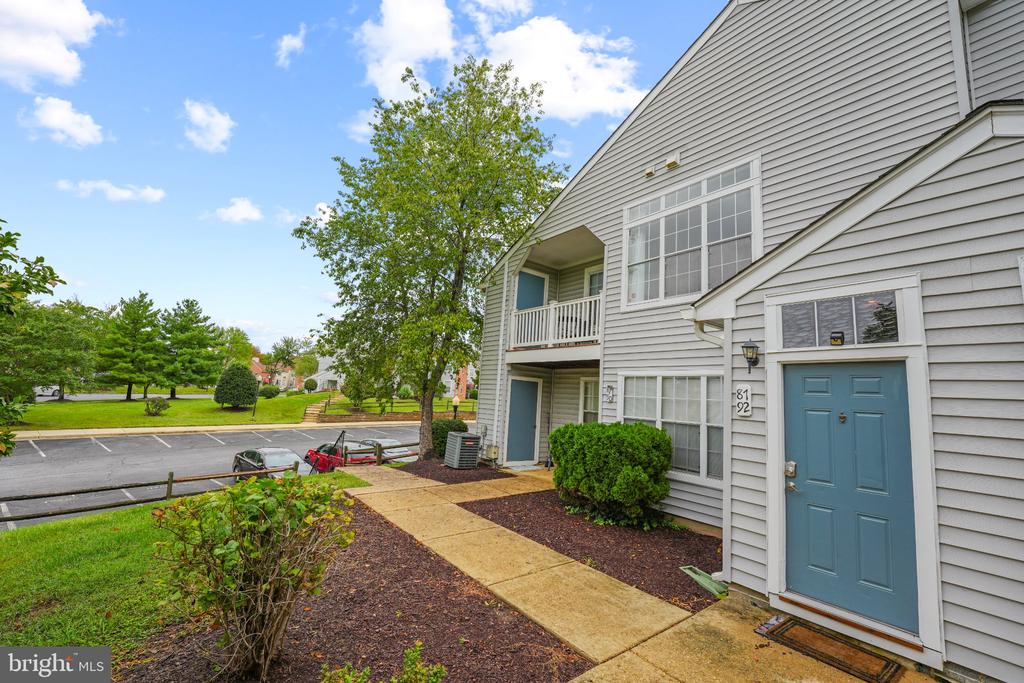 Gray-sided home entrance with a blue door, small balcony, and landscaped walkway beside a parking lot.