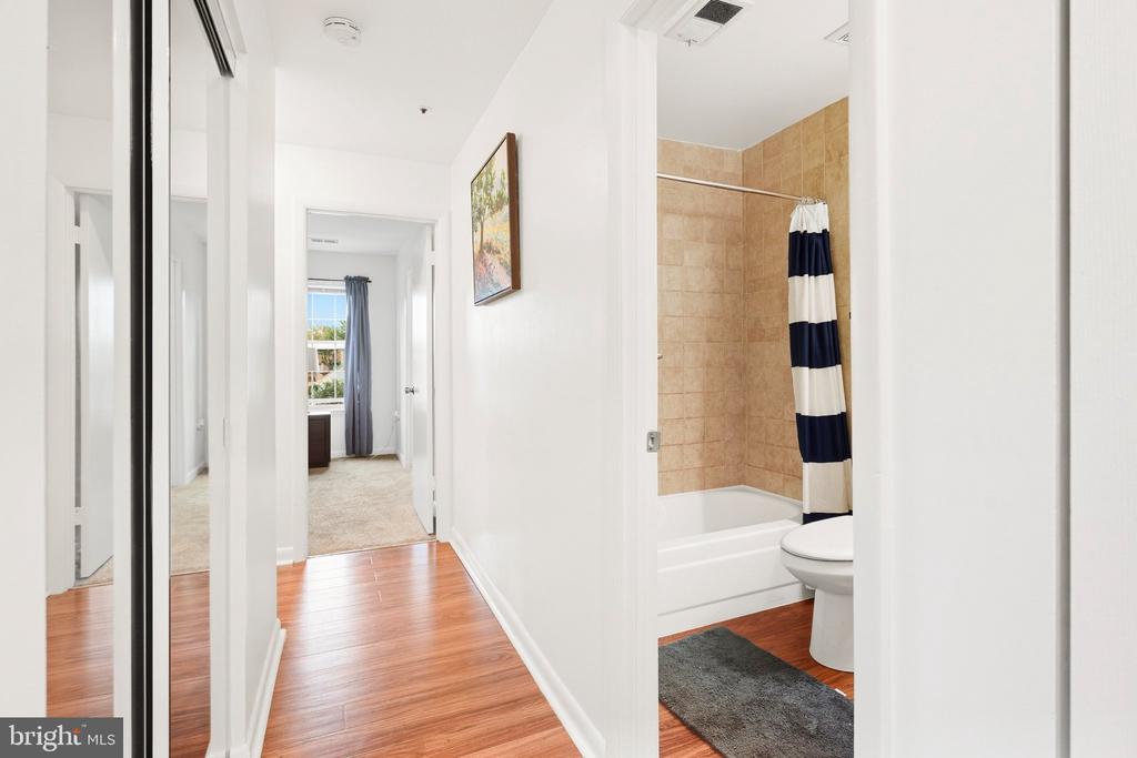 Bright hallway with wood flooring, mirrored closet doors, and an open bathroom with a striped shower curtain.