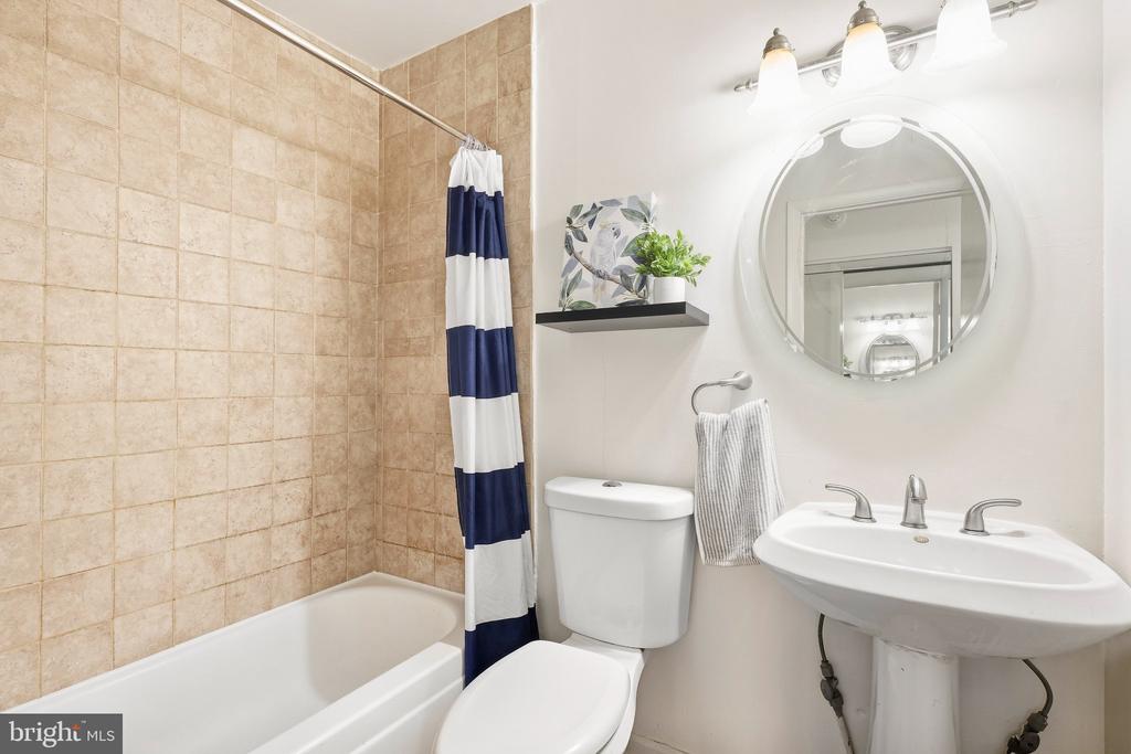 Bathroom with tan tiled tub surround, striped shower curtain, toilet, and pedestal sink beneath a round mirror.