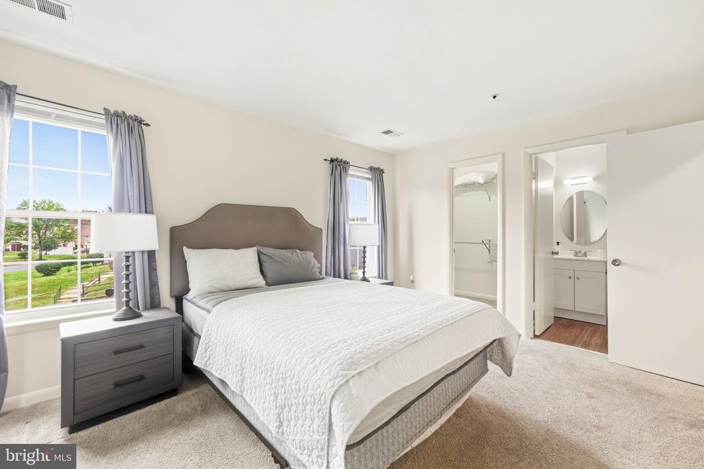 Staged bedroom with a large bed, gray headboard, two bright windows, and an adjoining bathroom.