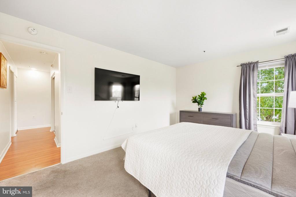Bedroom with a white quilted bed, wall-mounted television, and curtained window overlooking trees.