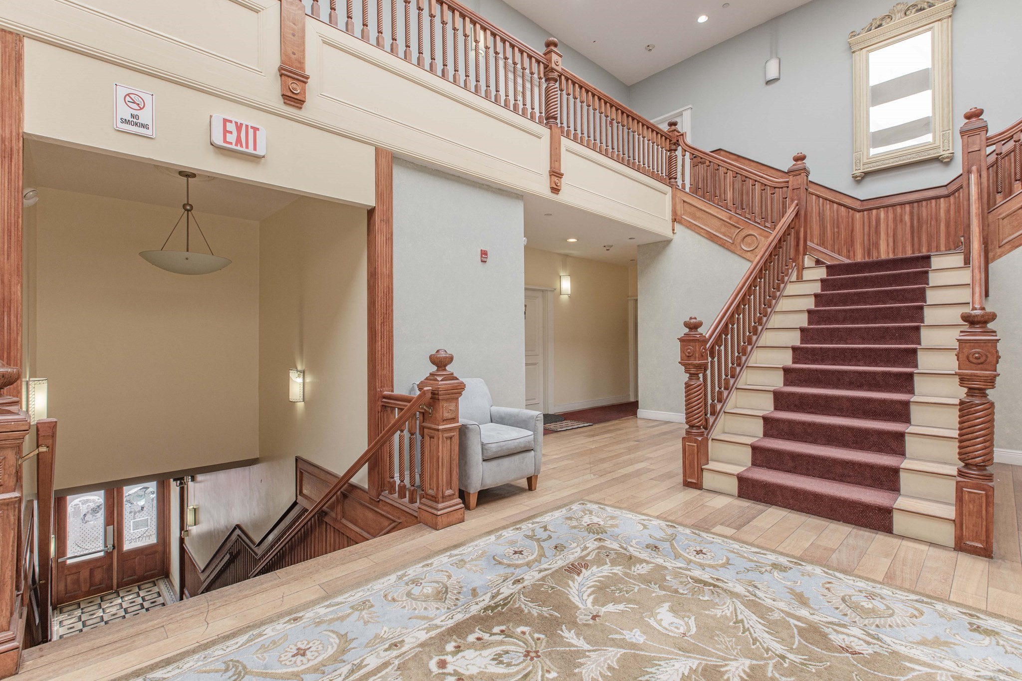 Interior lobby with an ornate wooden staircase, burgundy runner, and gray armchair.