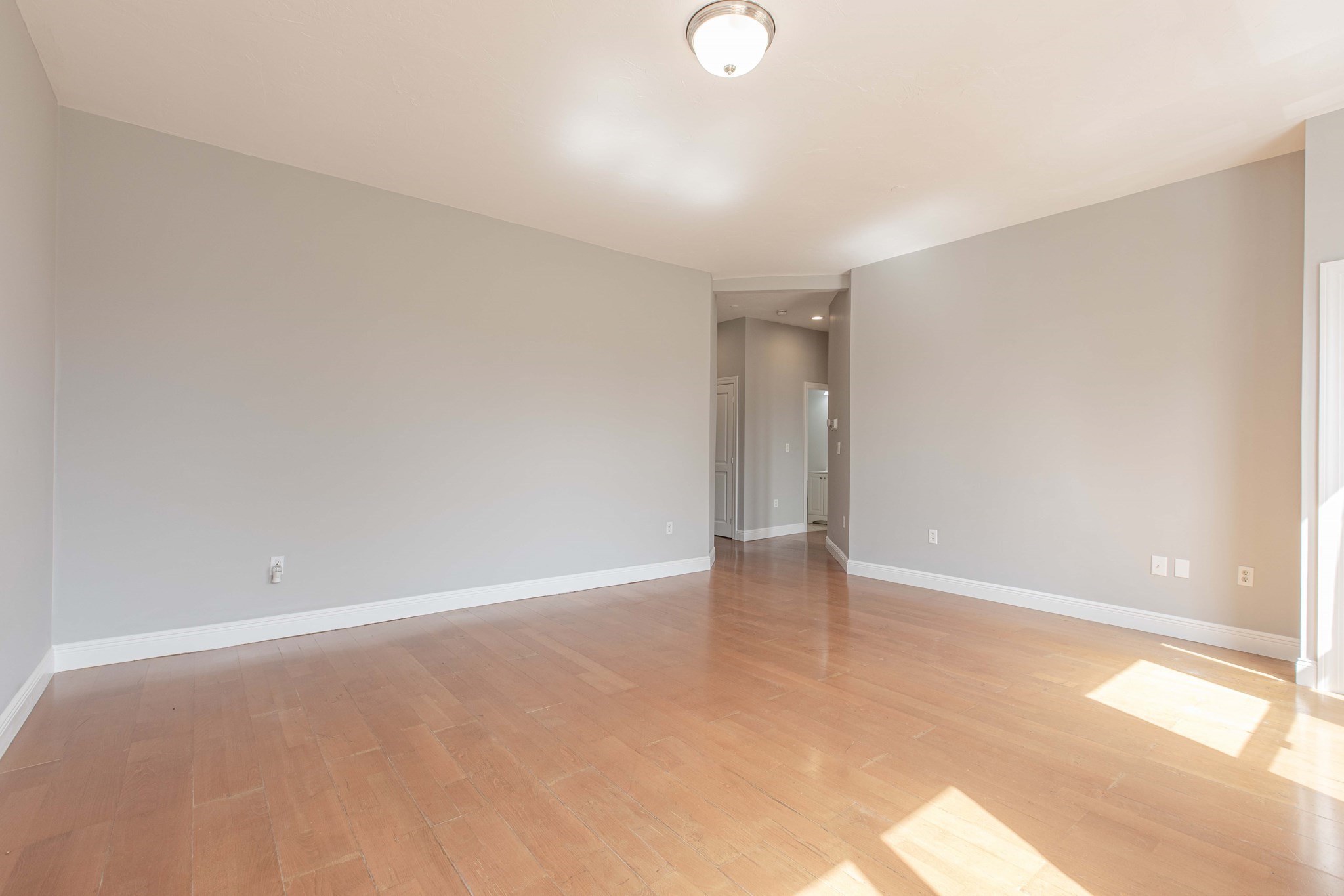 Empty living area with gray walls, reddish wood flooring, and sunlight on the floor.