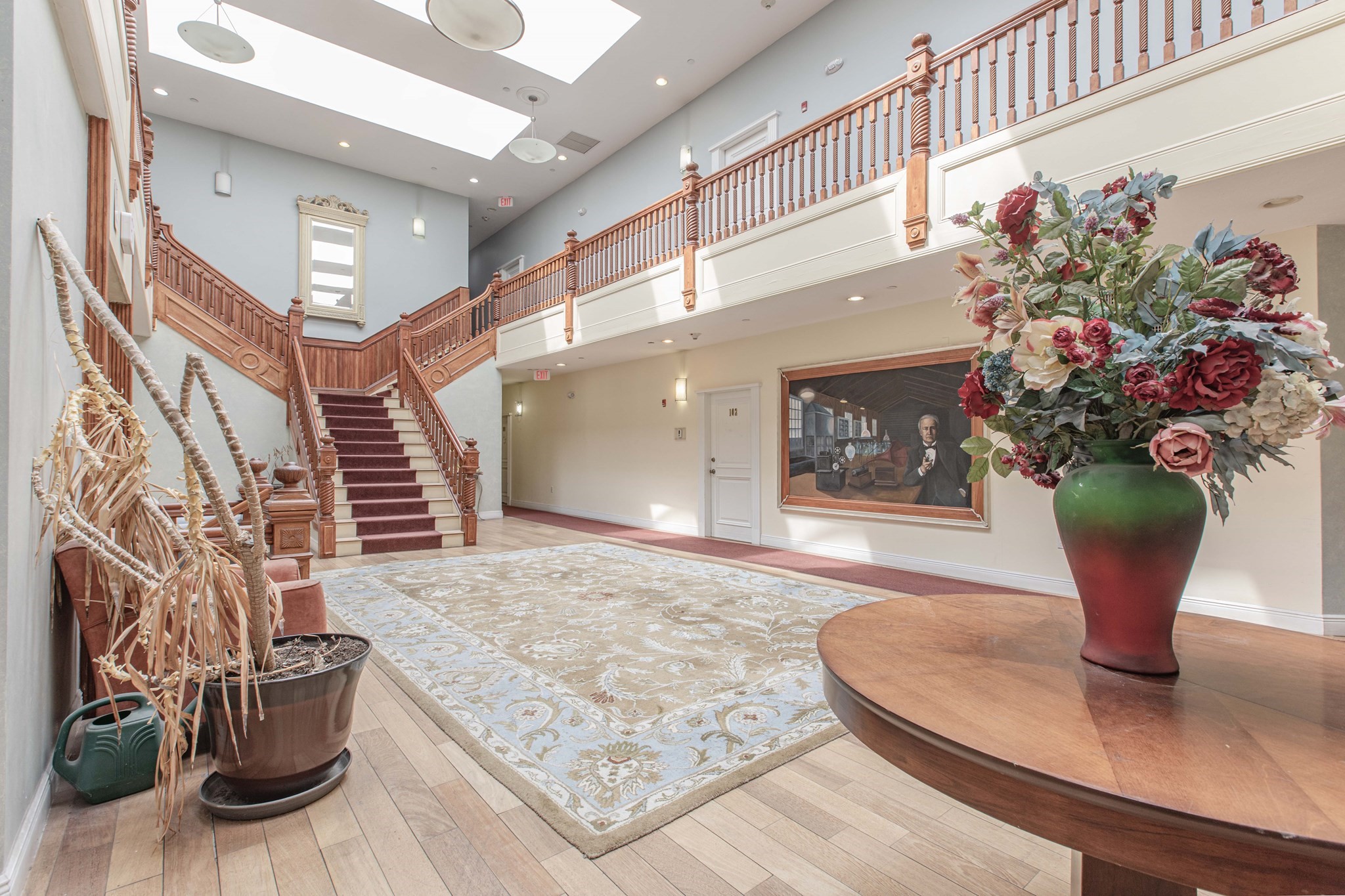 Building lobby with a grand wooden staircase, upper balcony, skylight, and floral arrangement.