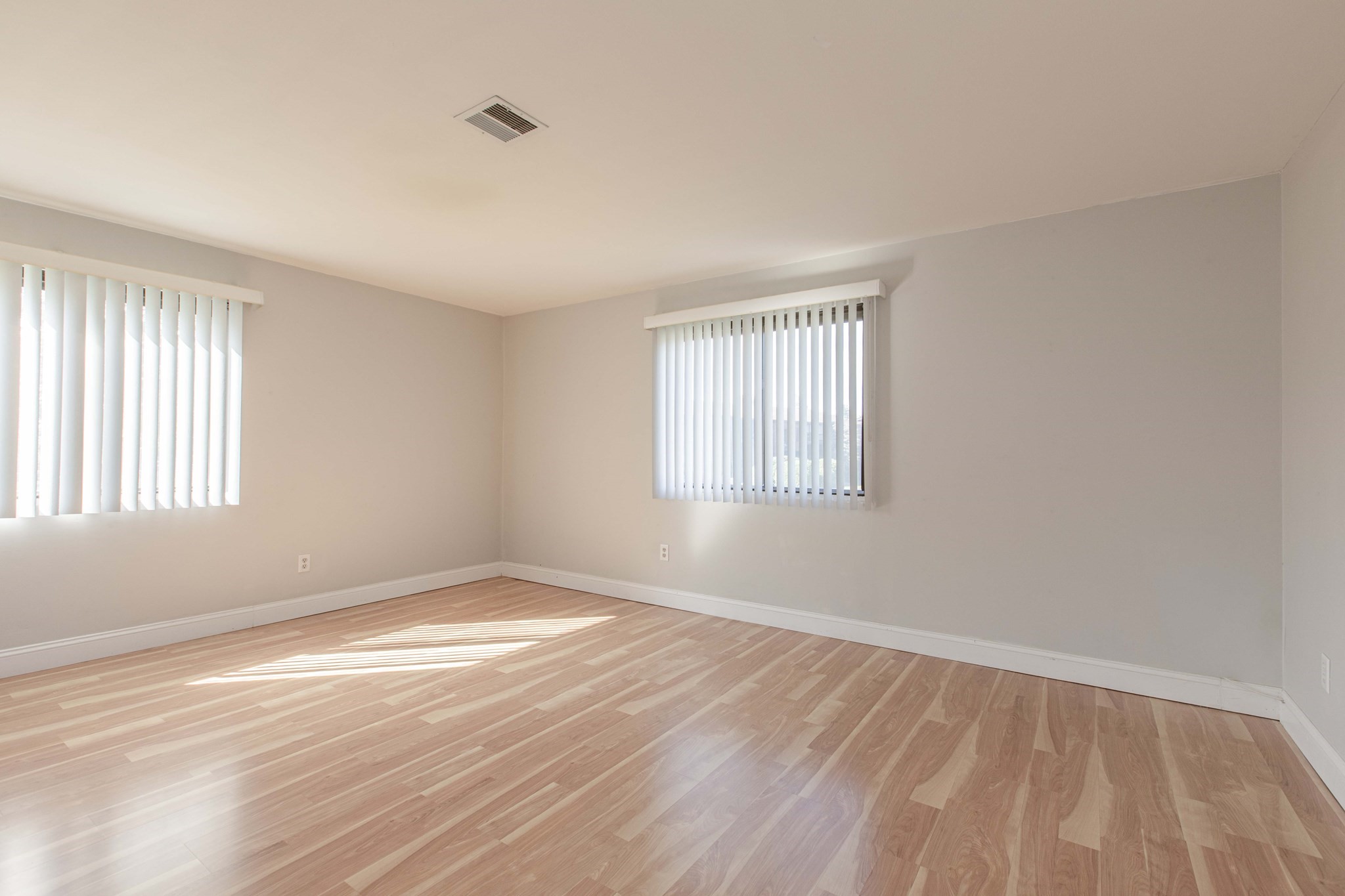Empty room with light wood flooring, two windows with vertical blinds, and sunlight across the floor.