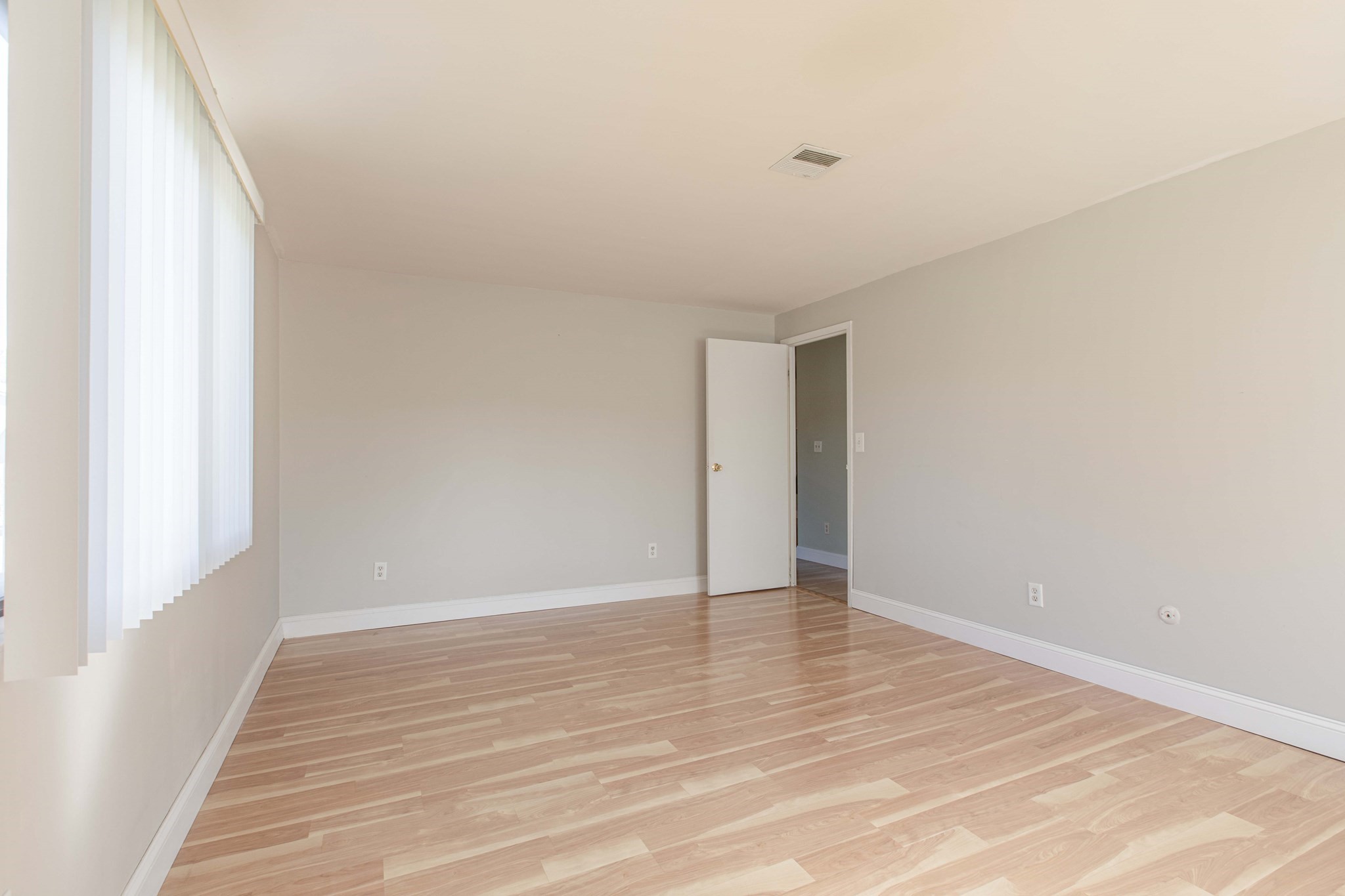 Vacant room with light wood-look floors, tall windows with vertical blinds, and an open doorway.