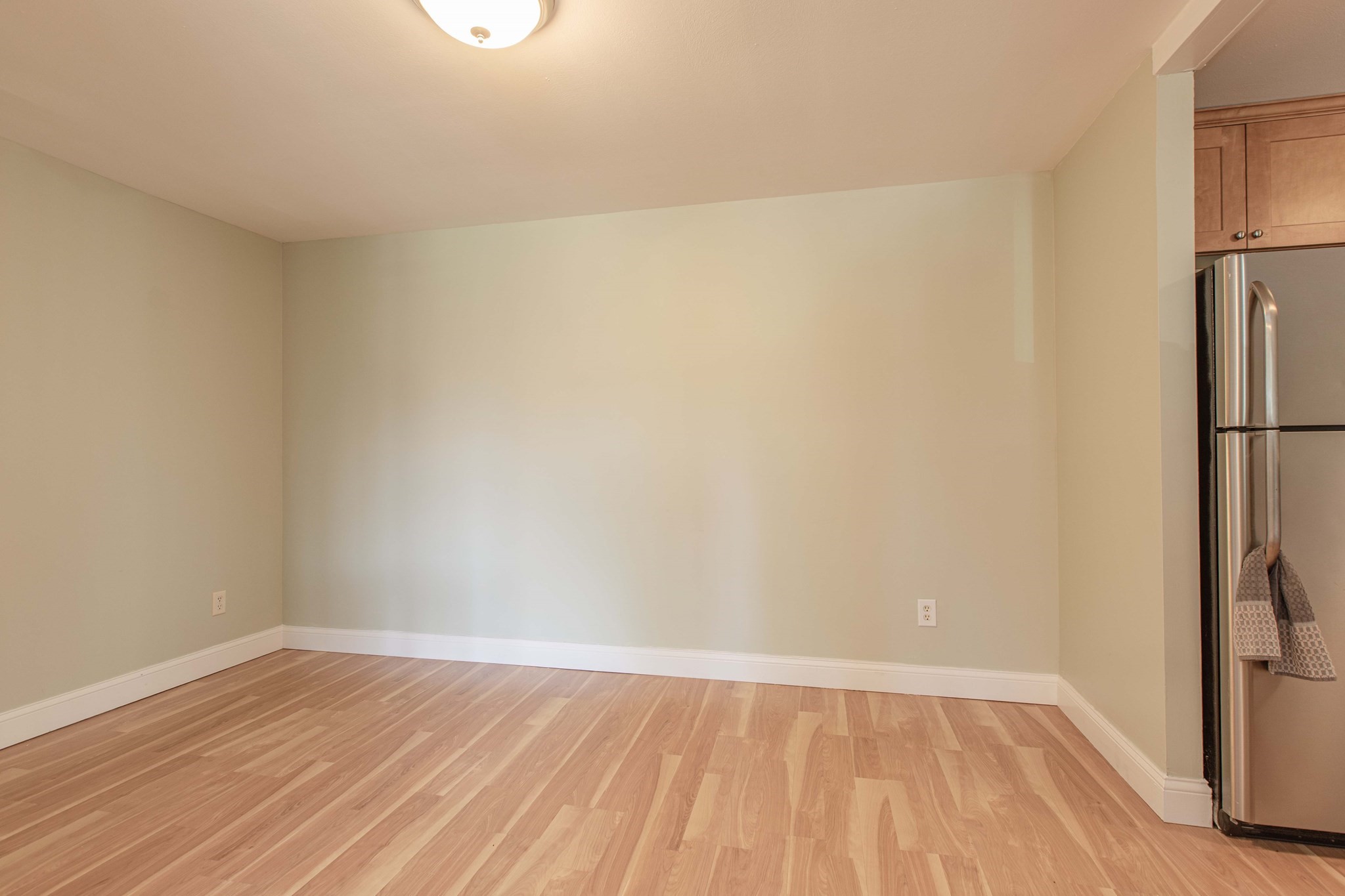 Empty apartment room with light wood flooring beside wooden kitchen cabinets and a stainless-steel refrigerator.