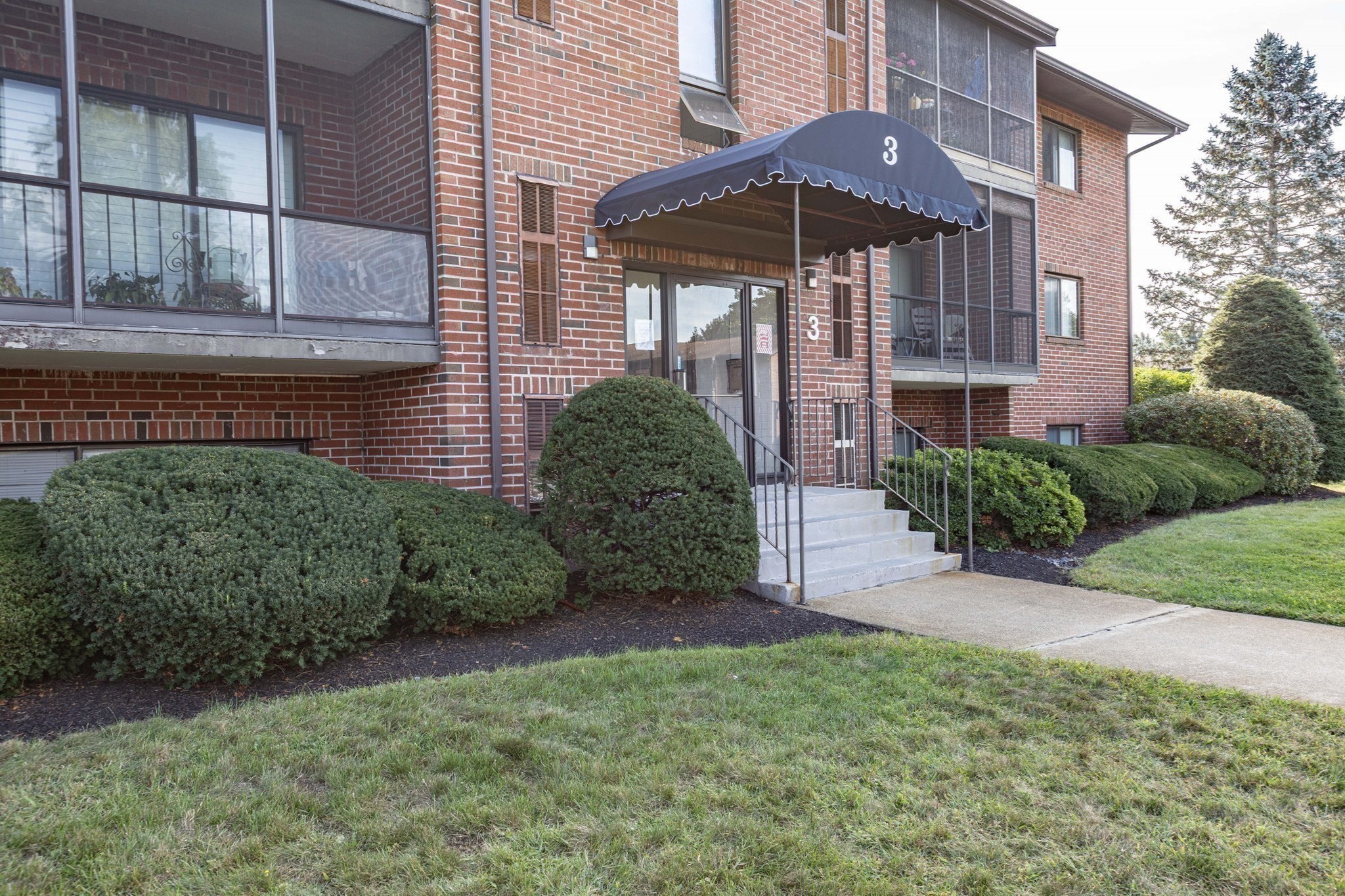 Red-brick building entrance with a number 3 awning, concrete steps, and landscaped shrubs.