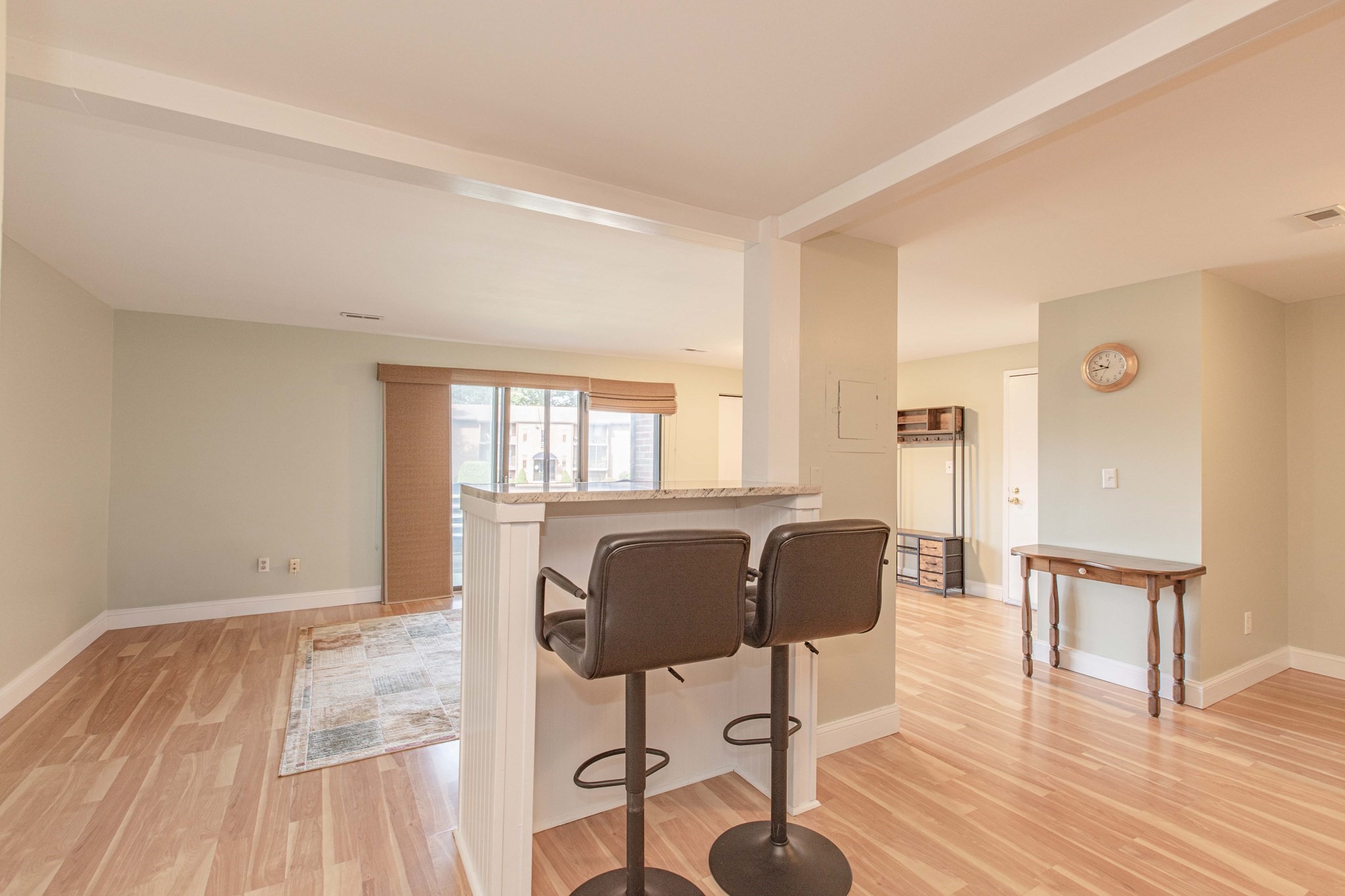 Open-plan living area with a breakfast bar, two stools, light wood floors, and a large shaded window.