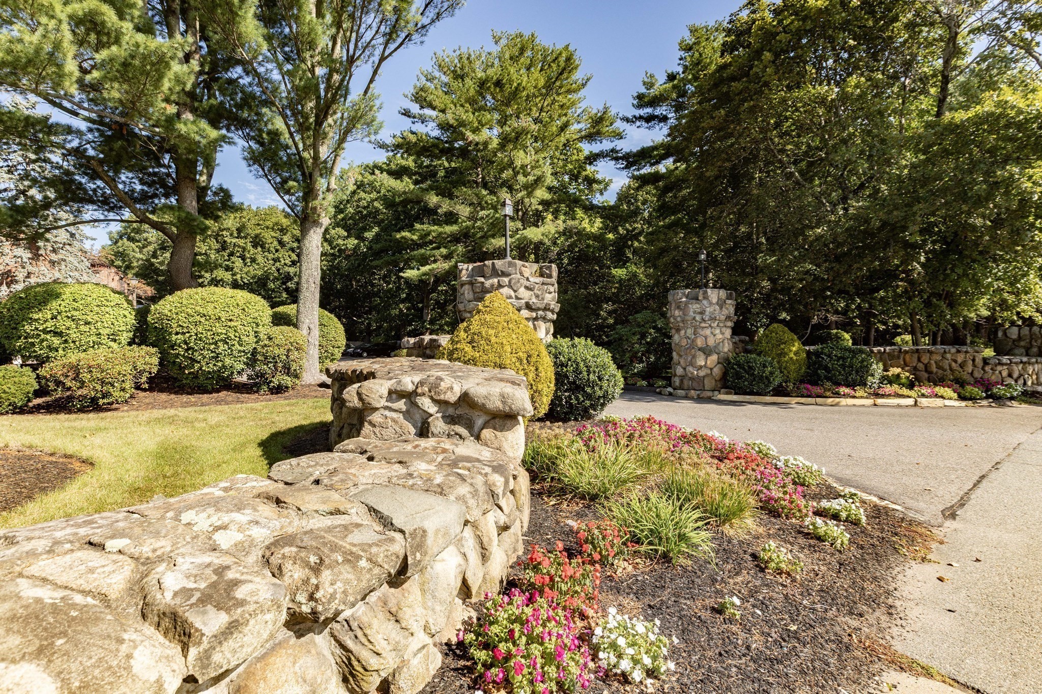 Landscaped entrance with stone walls and pillars, mature trees, and colorful flower beds beside a paved drive.